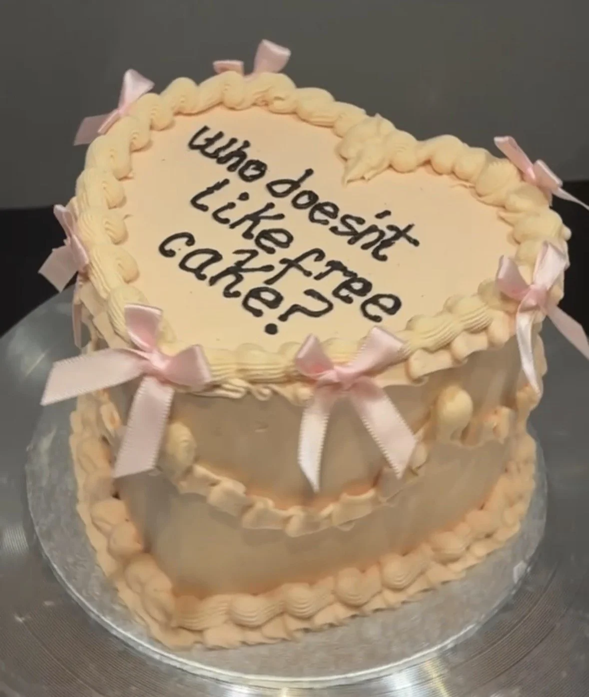 A heart-shaped cake with pink ribbons and cream borders, with the message in black icing: 'Who doesn't like free cake?'