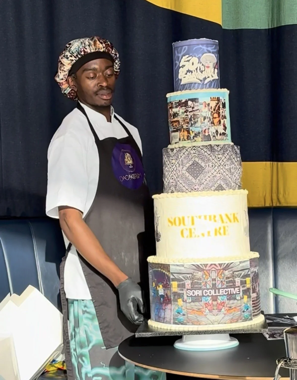 Zhora wearing a chef's uniform, apron, and a colorful bonnet standing next to a tall, multi-tiered cake with various images and logos. The background has dark curtains with yellow and green accents.