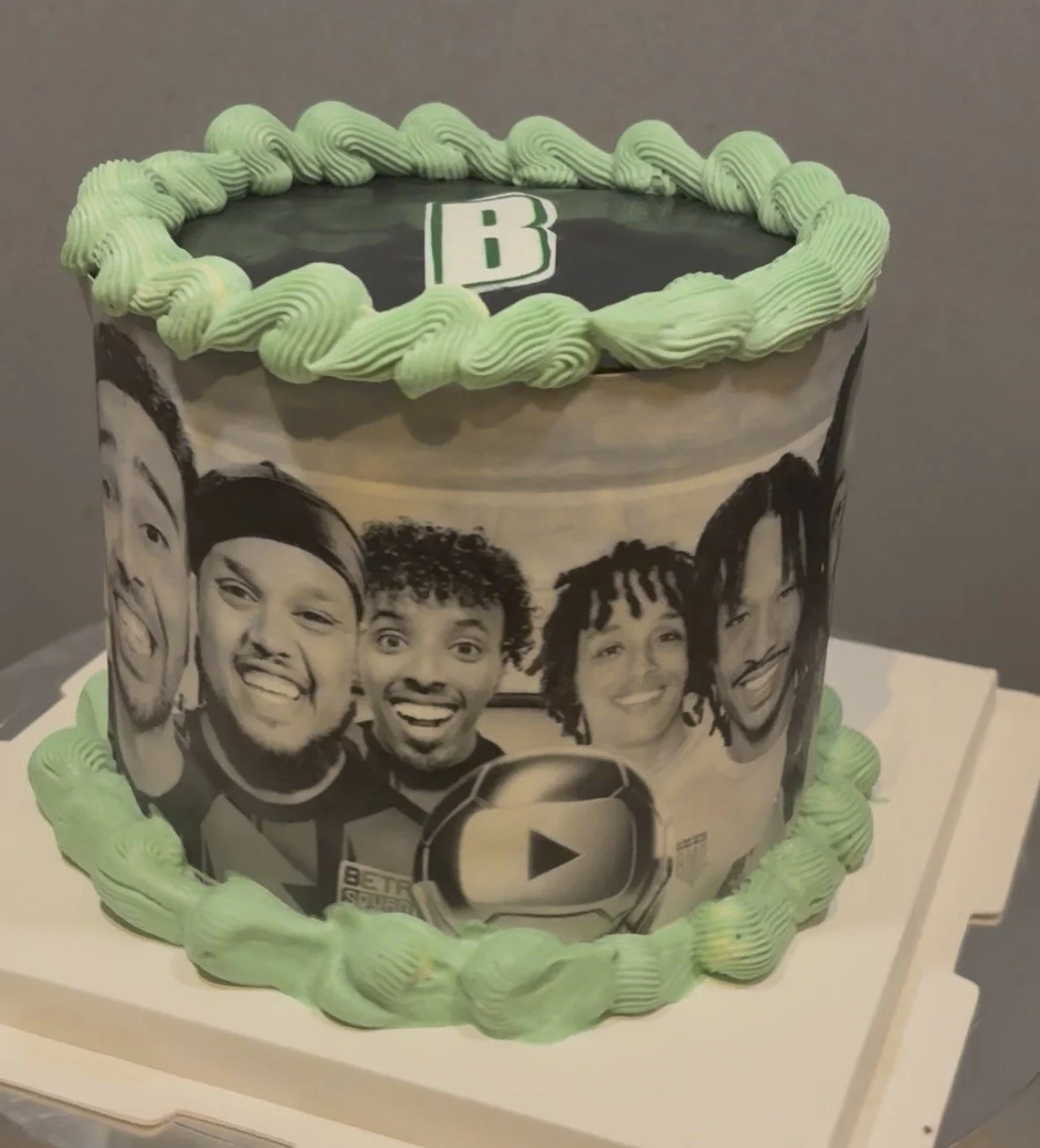 A decorated cake with a black and white photo of four smiling people on the side and a green and black top with a letter B and a green piped border.