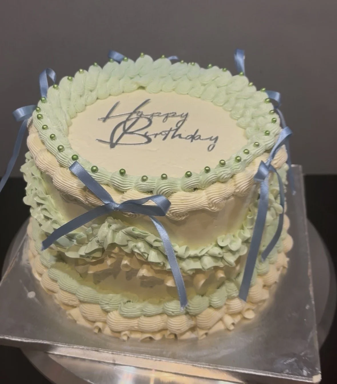 A two-layer birthday cake decorated with white and green frosting, blue ribbons, and small green pearls. The top has a white circular top with the words 'Happy Birthday' written in black.