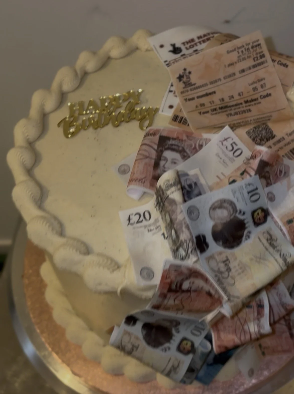 A birthday cake with white icing and a gold "Happy Birthday" topper, decorated with various UK banknotes and lottery tickets.
