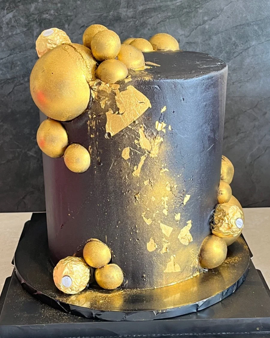 A black cylindrical cake decorated with gold spheres and gold leaf accents, placed on a black base.