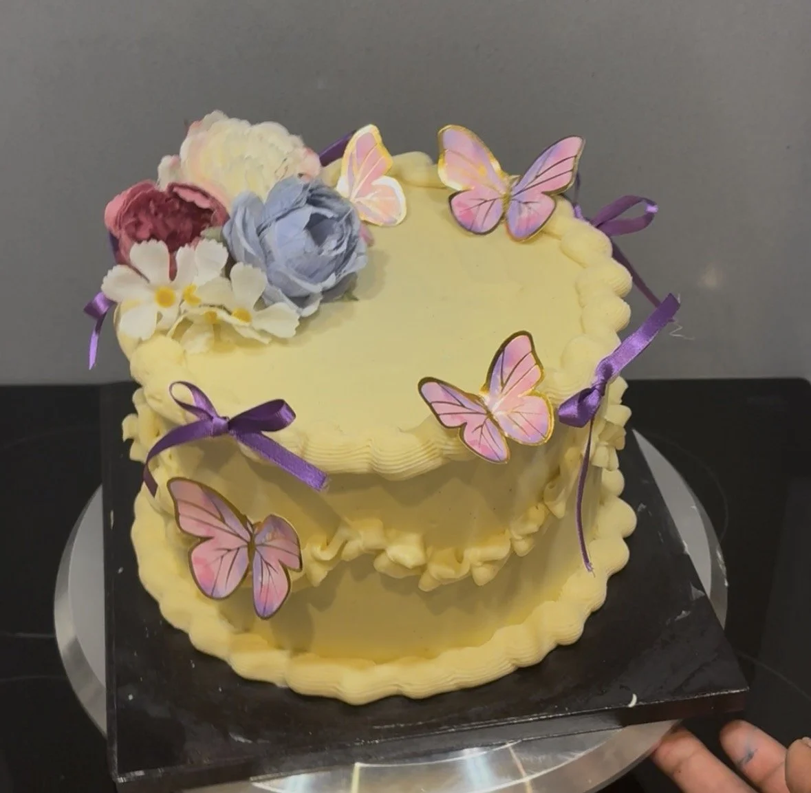 Yellow two-tiered cake decorated with purple ribbons, pink and purple flowers, and pink butterfly toppers.