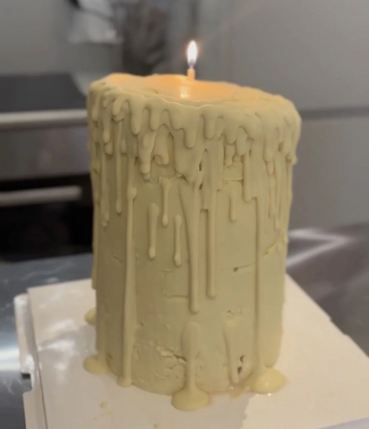A white frosted birthday cake with melted white chocolate dripping down the sides and a single lit candle on top.