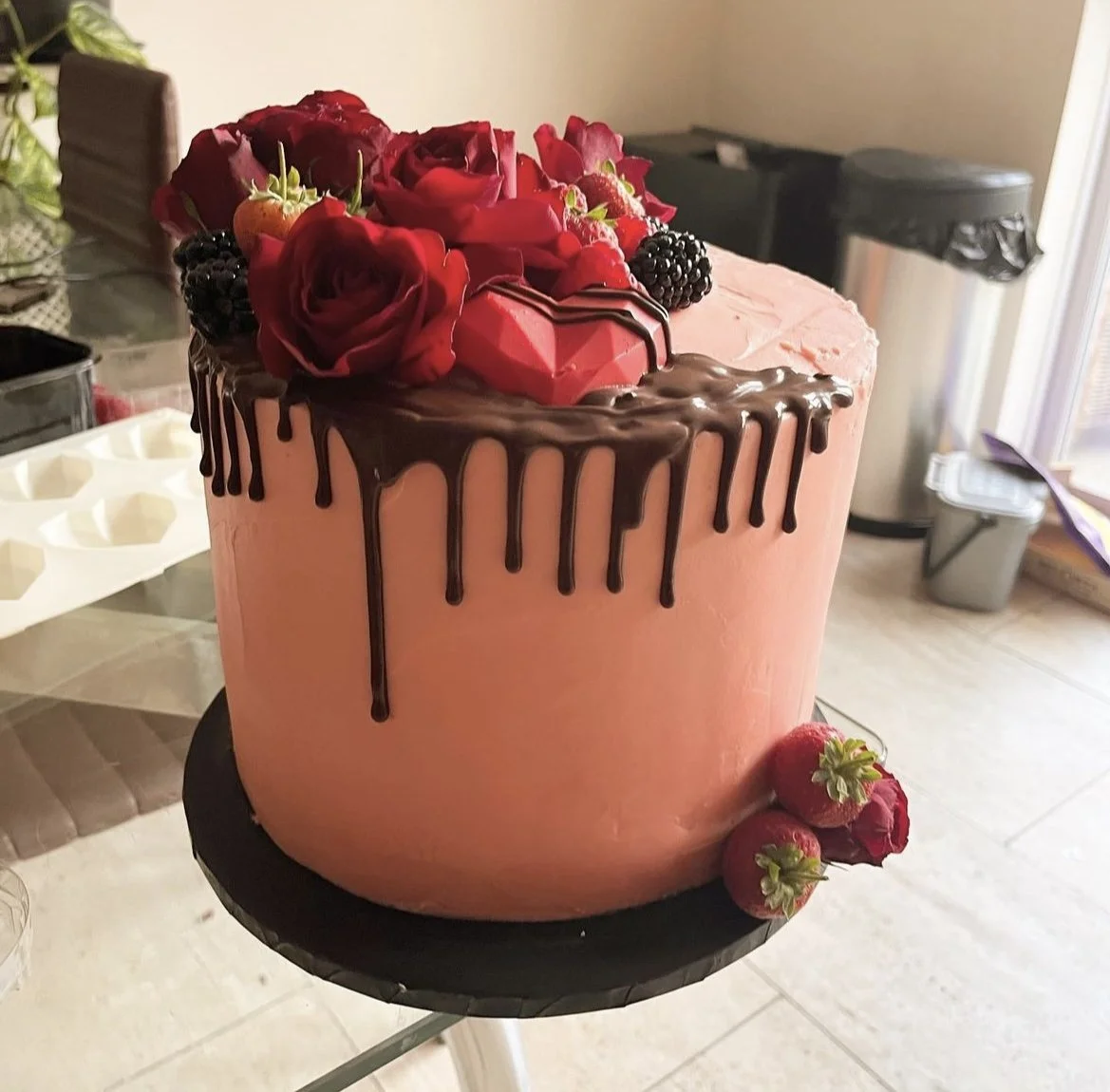 A pink cake with chocolate dripping from the top edge, decorated with red roses, strawberries, blackberries, and a pink heart-shaped decoration on top, sitting on a black cake stand.