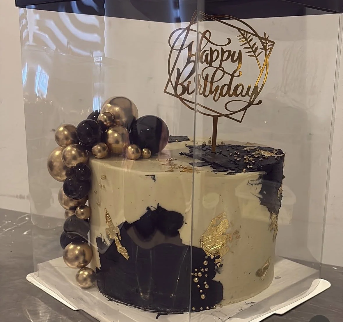 Decorated birthday cake with black, white, and gold accents, topped with a "Happy Birthday" cake topper, inside a clear display case.