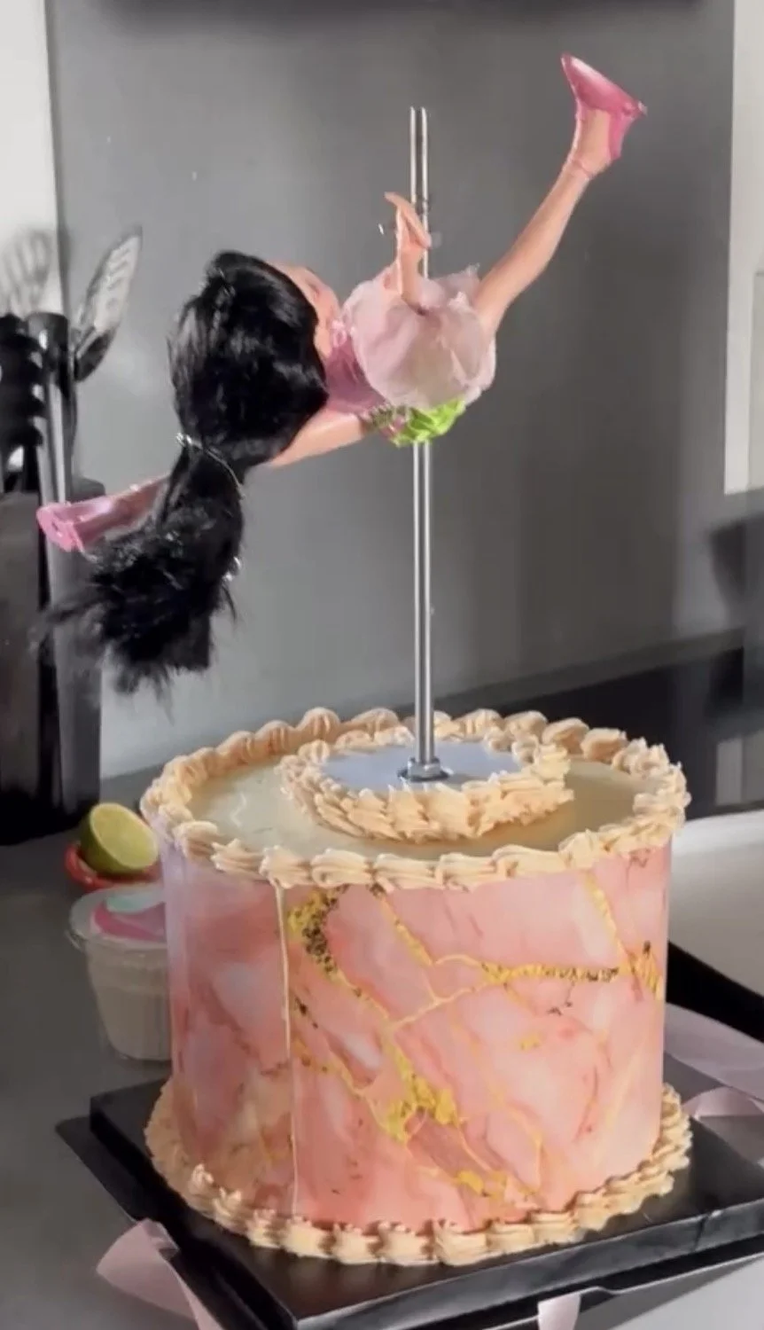 A large round pink cake with marbled gold streaks and decorative piping on top and around the edges, featuring a ballerina figurine on a pole in the center, with a lime half on the side of the cake.