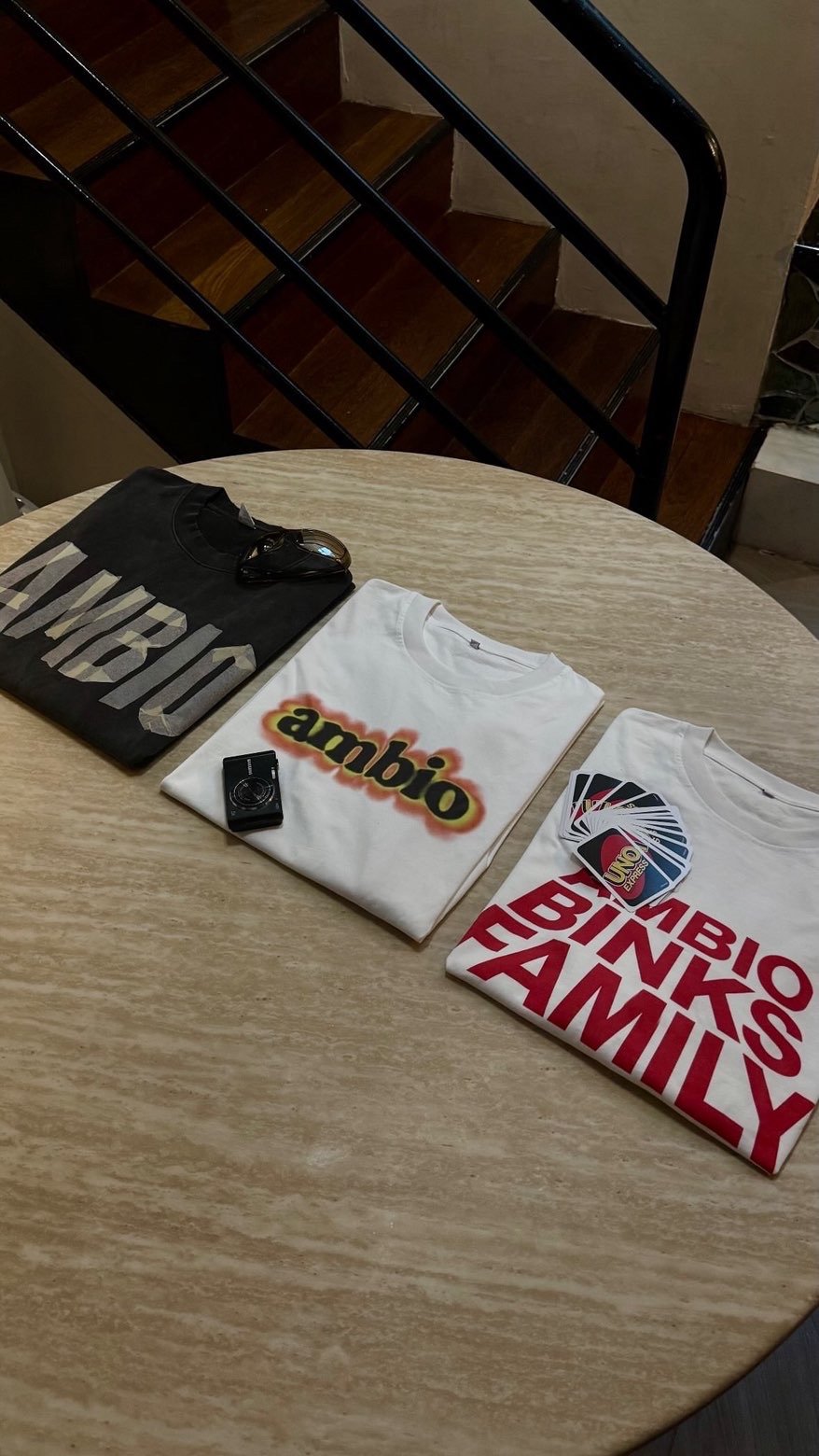 Trois t-shirts pliés avec des logos et des cartes de jeu sur une table ronde en bois, en arrière-plan un escalier en bois avec une rampe métallique.