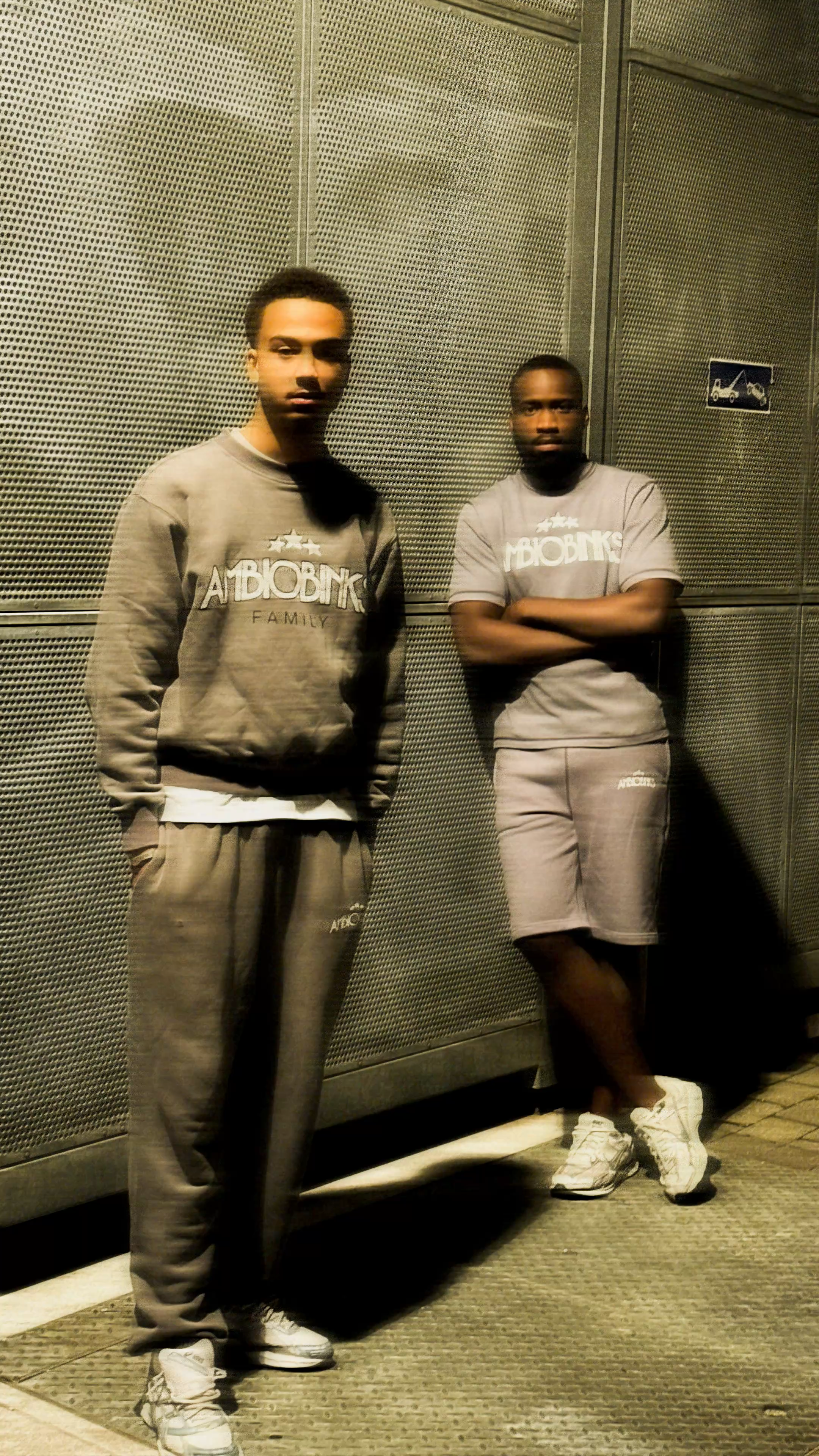 Deux jeunes hommes debout contre un mur en métal perforé, l'un portant un sweat gris avec le mot 'AMBIBIK' et l'autre un t-shirt gris avec le même logo, tous deux en vêtements décontractés.