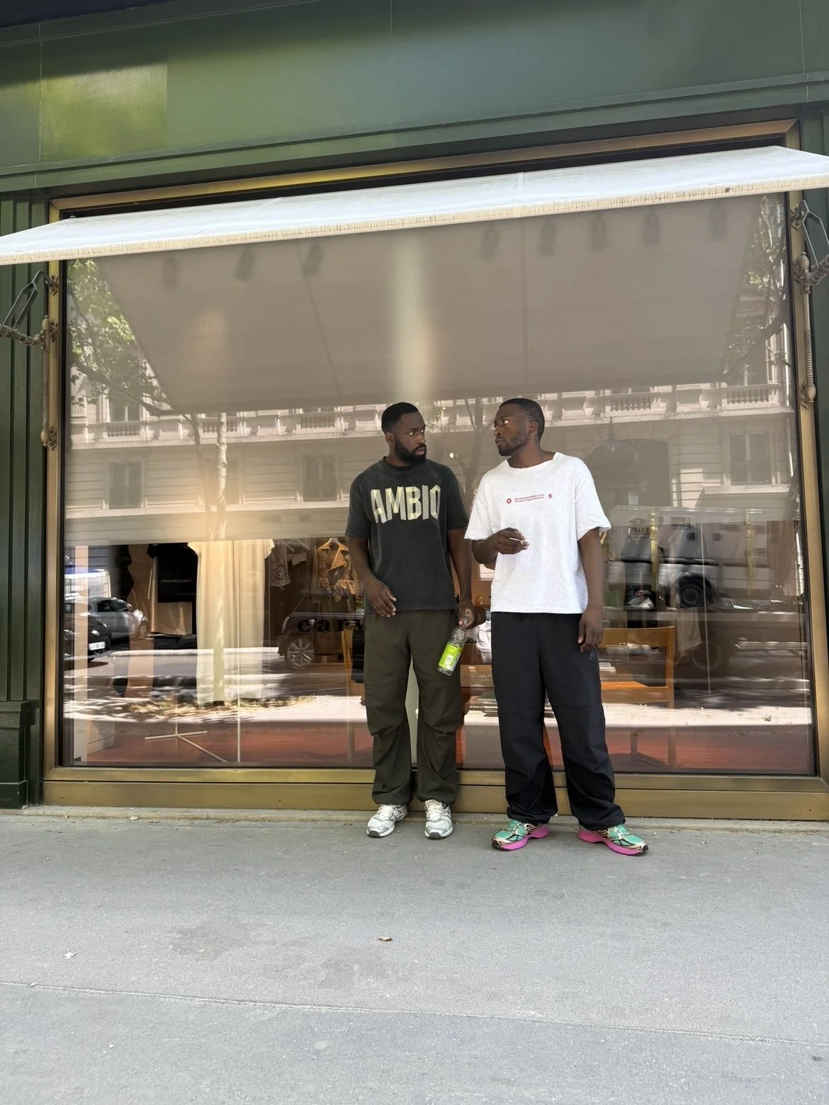 Deux hommes discutent devant une vitrine d'un magasin, l'un porte un t-shirt noir avec 'AMBIO' écrit dessus et l'autre un t-shirt blanc, portant un bouteille d'eau verte.