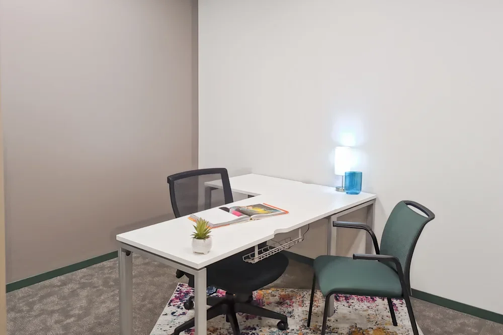 An empty office corner with a white desk, two chairs, a potted succulent, a magazine, and a small lamp on the desk.