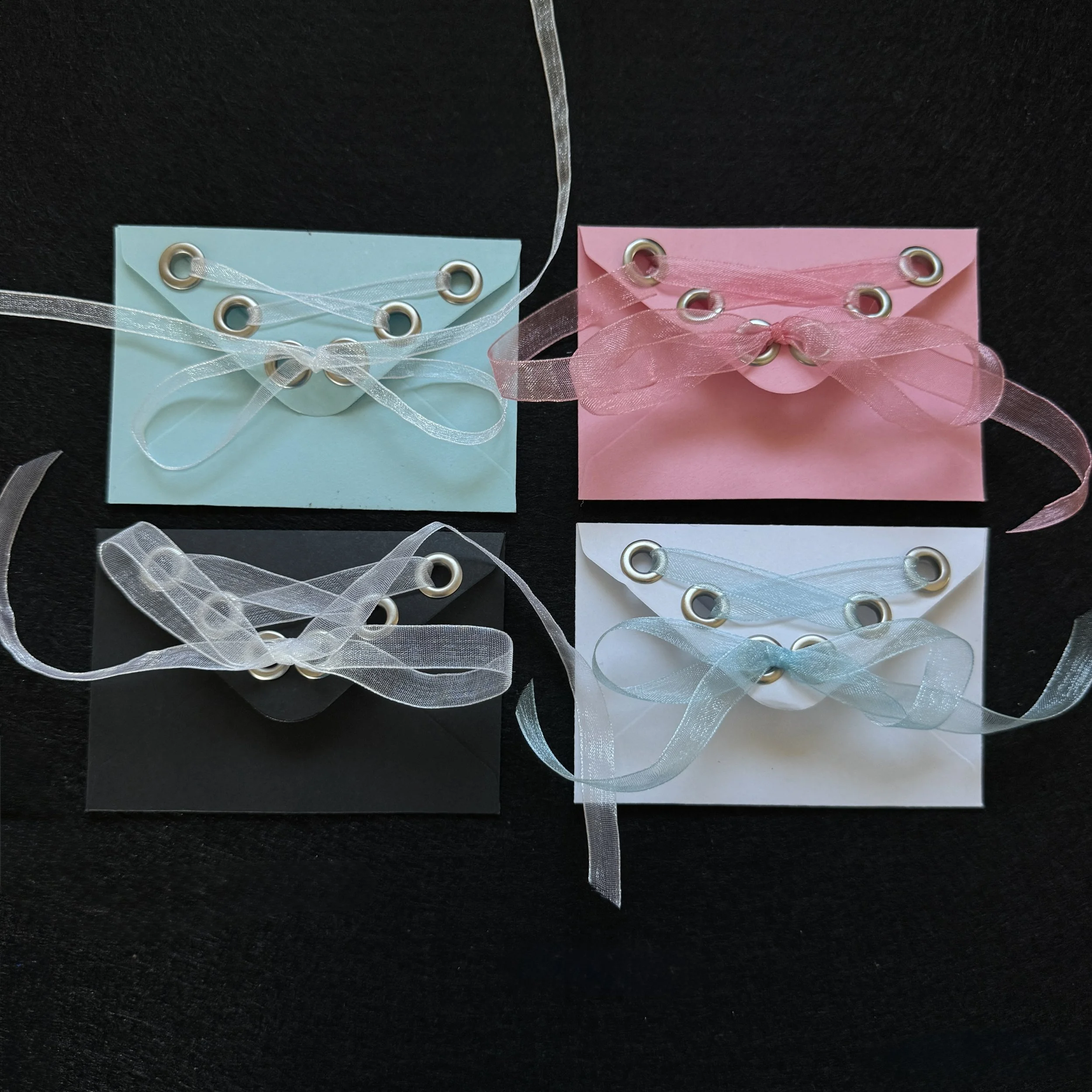 Four small envelope cards, two light blue and two pink, each decorated with small silver eyelets and tied with sheer ribbons, displayed on a black surface.