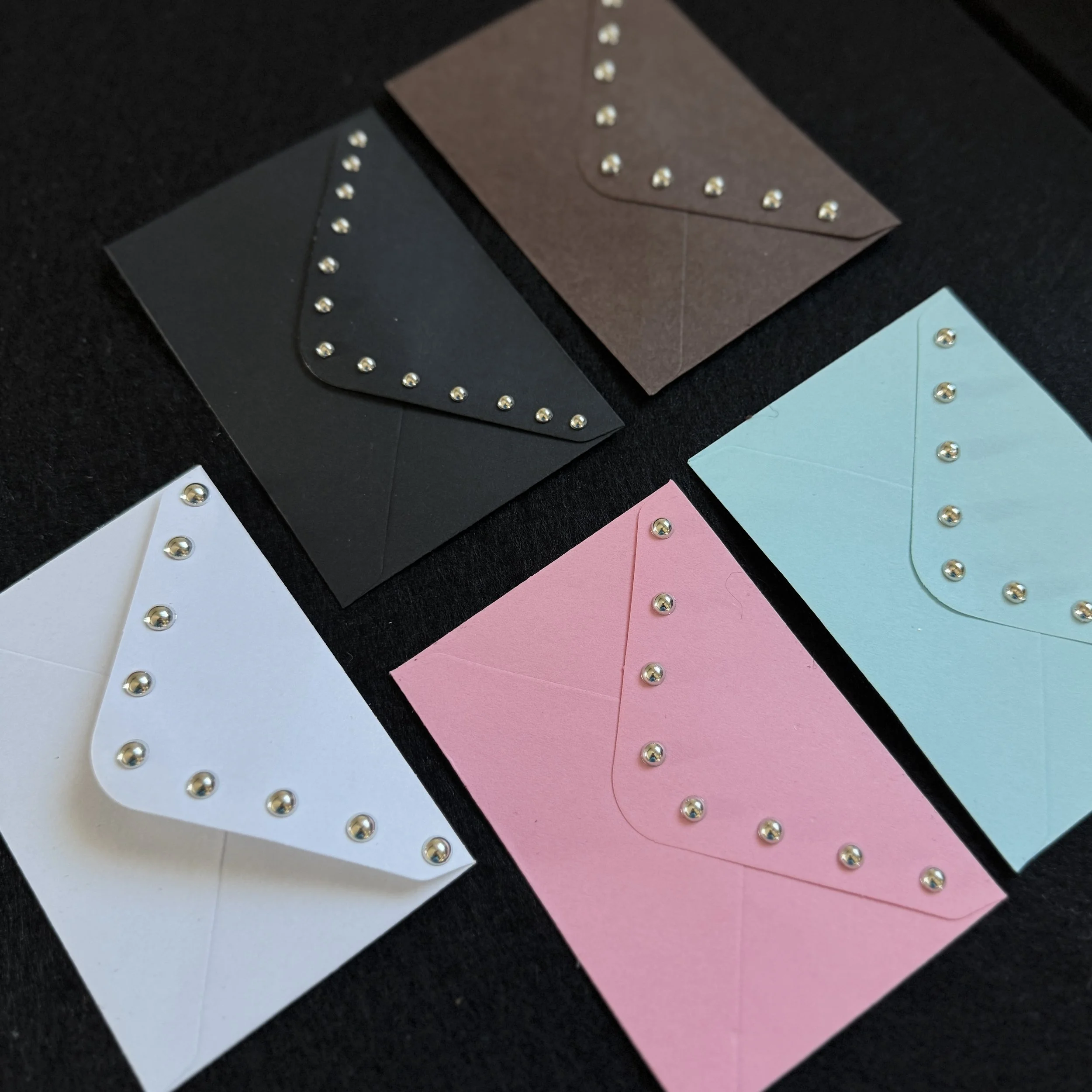 Close-up of several folded paper envelopes in white, pink, blue, black, and brown colors, decorated with small gold-colored embellishments around the edges, placed on a black surface.