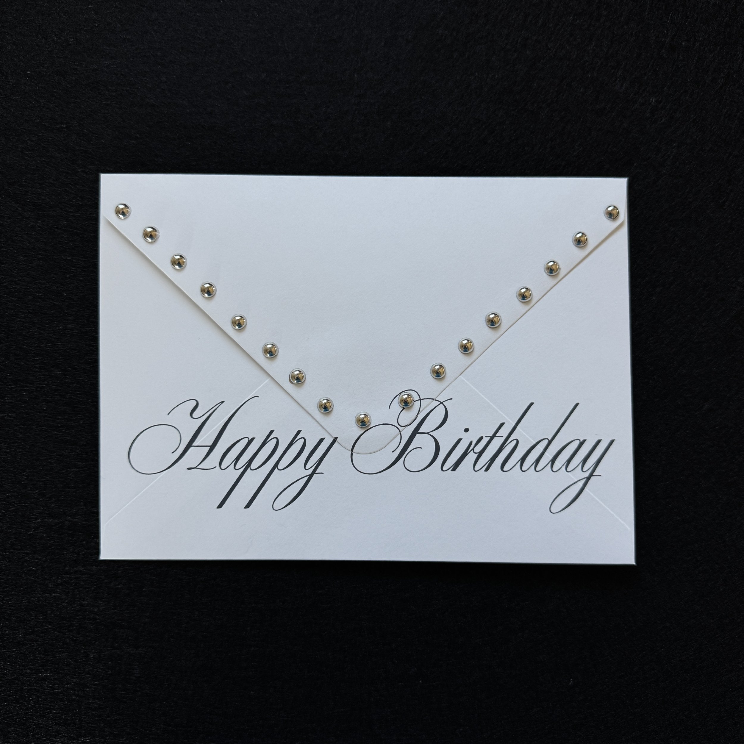 Close-up of a white birthday card with the words "Happy Birthday" written in elegant black cursive, decorated with small metallic silver studs on a black textured background.