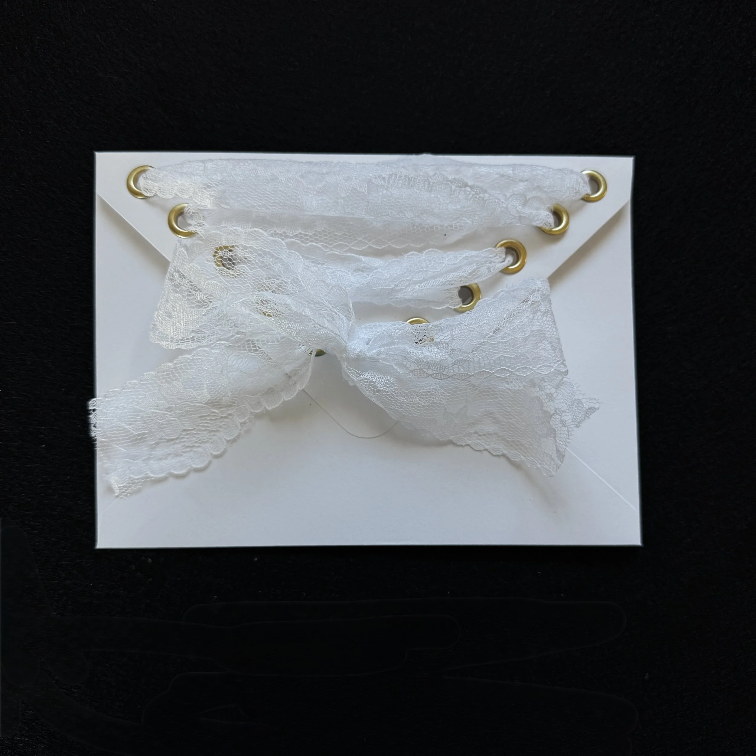 White lace fabric with gold eyelets placed on top of a white rectangular paper, set against a black background.