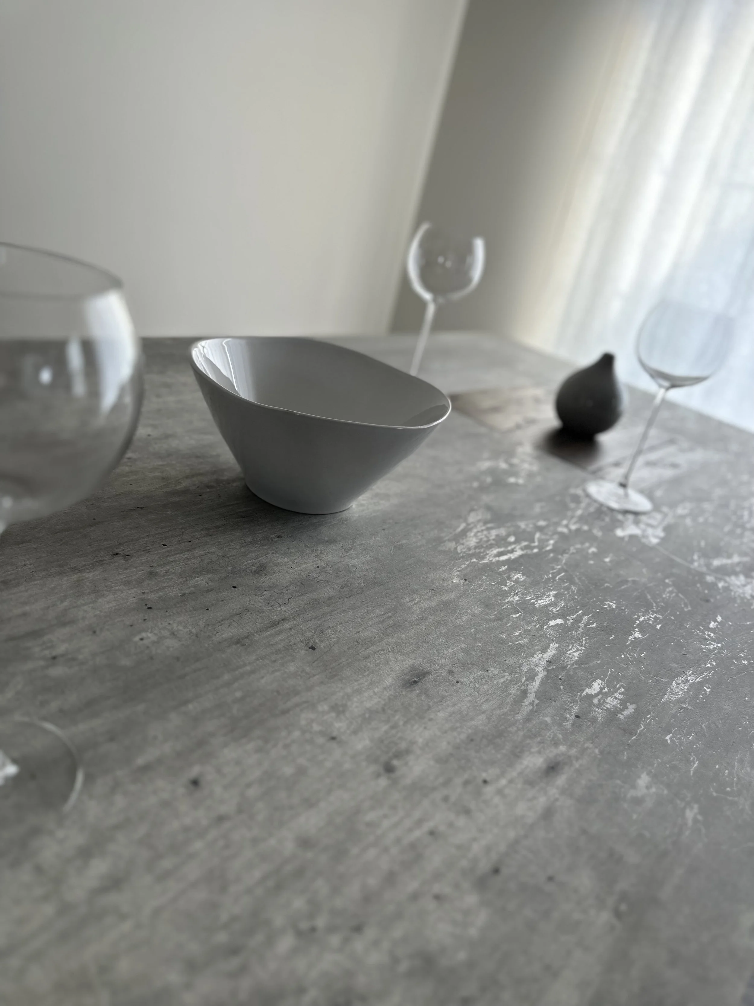 A close-up view of a textured gray surface with several glass tableware pieces, including a white bowl, two empty wine glasses, a black pear-shaped object, and a blurred background with a light curtain.