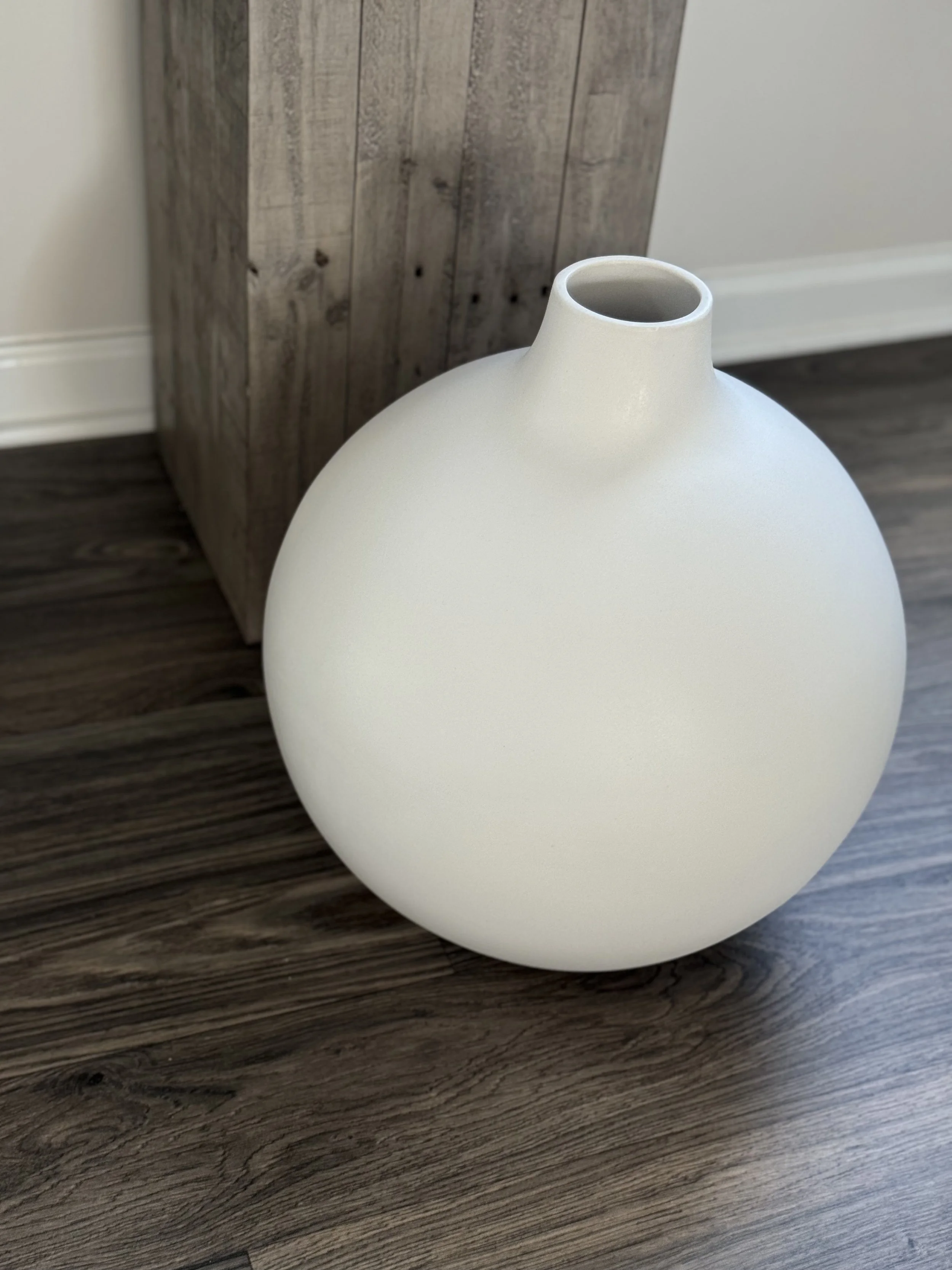 A white, round ceramic vase with a narrow opening at the top, placed on a dark wooden floor next to a wooden furniture piece and a white wall.