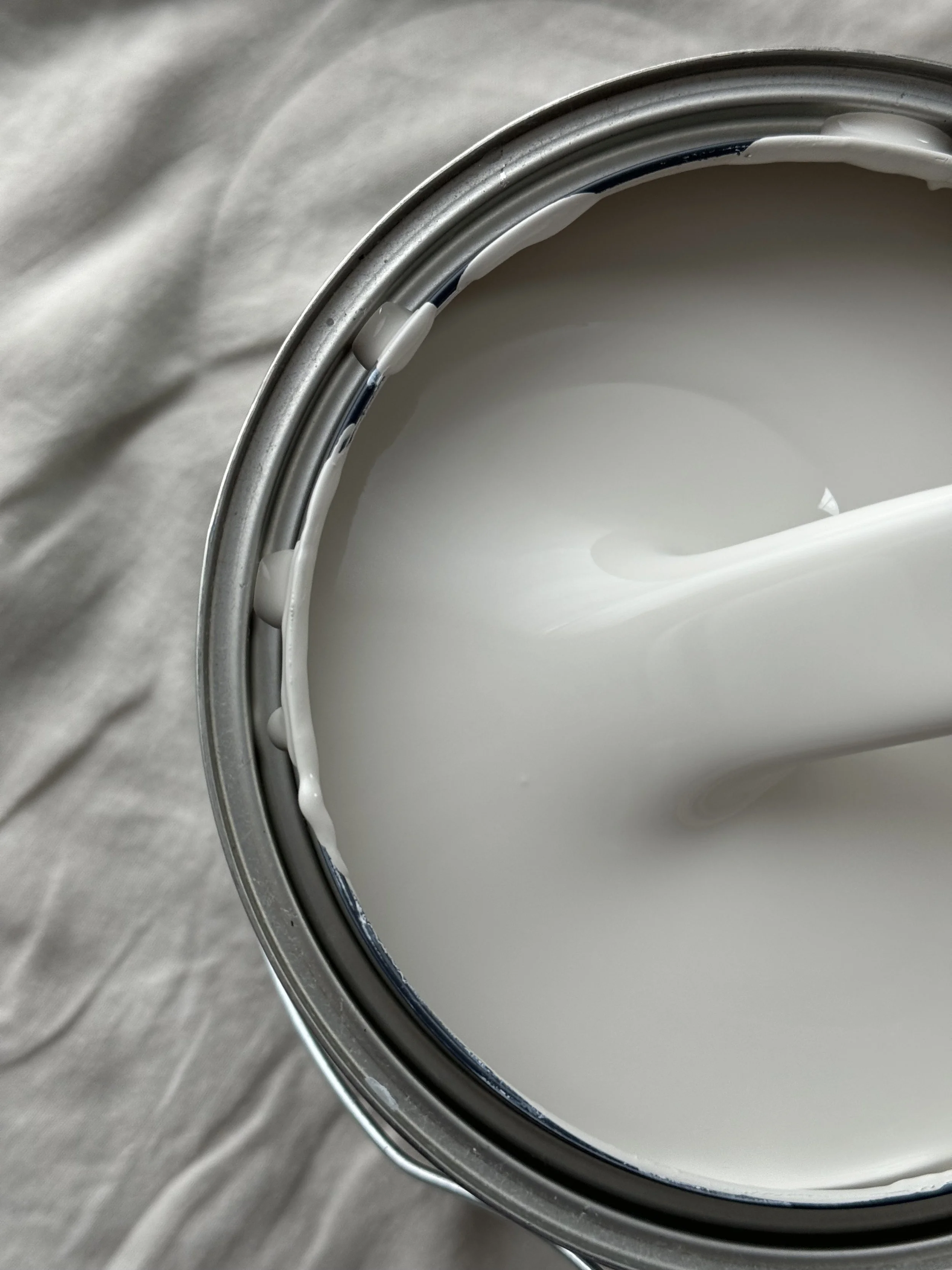 Close-up view of a can of white paint with a paintbrush dipped in it, on a light-colored wooden surface.