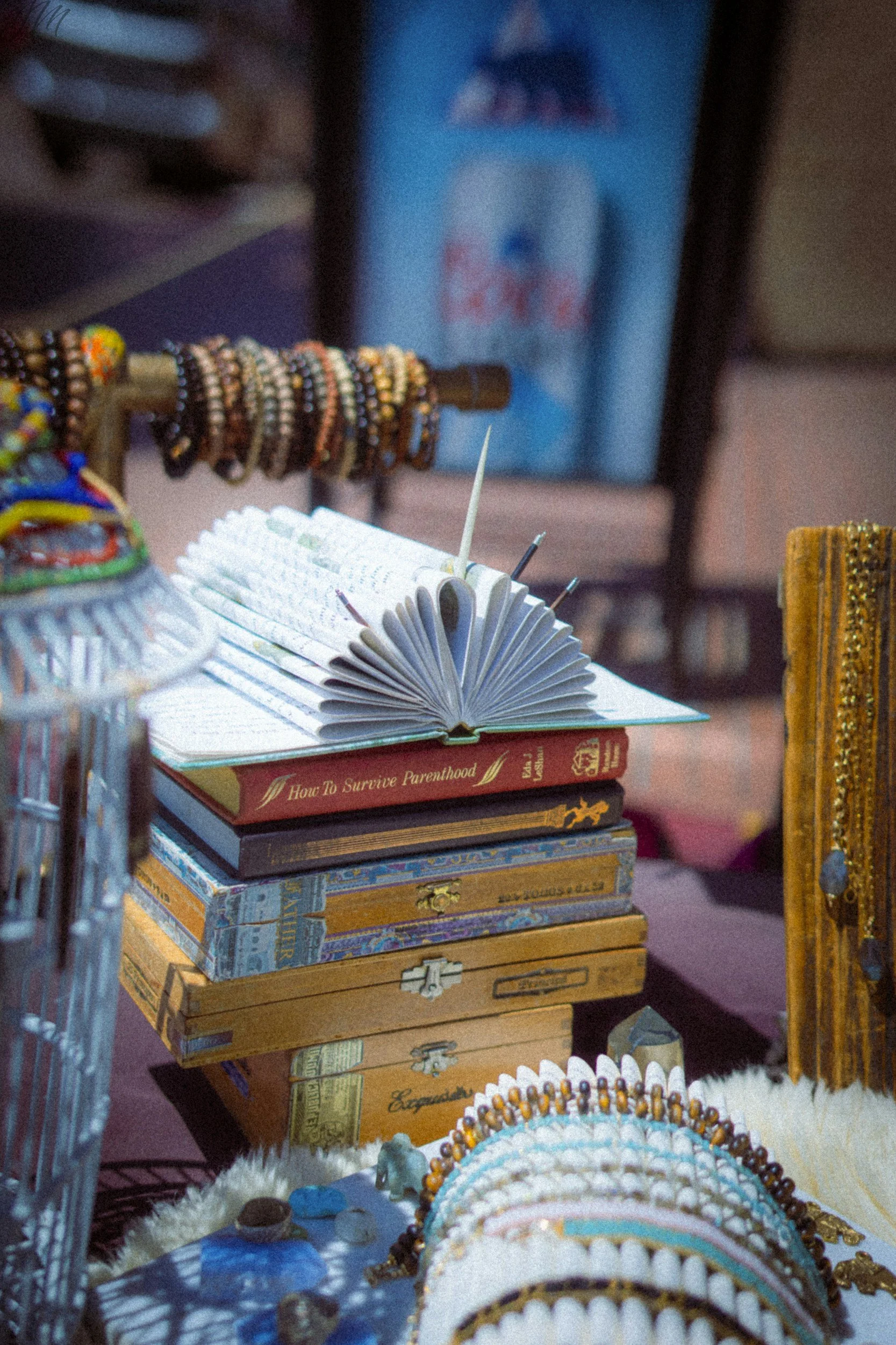 A table displays jewelry including earrings and bead necklaces, along with a stack of books, and a background with a blurry book cover and a rack holding bracelets.