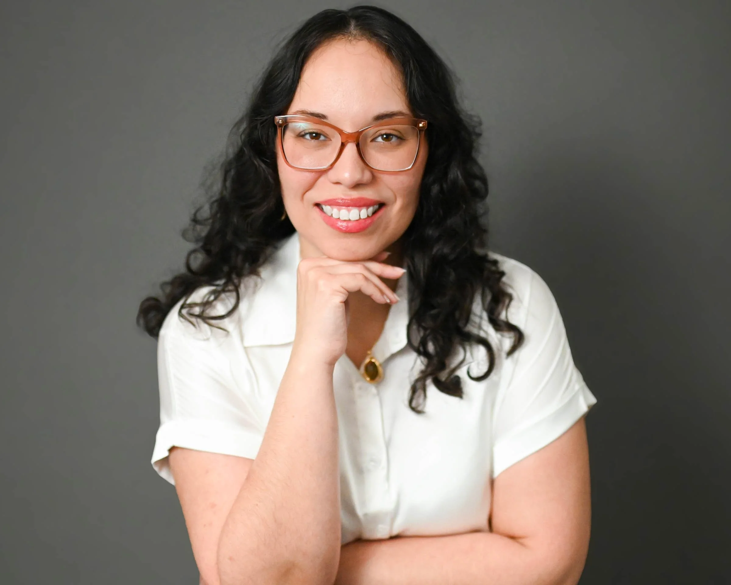A woman with long dark curly hair and glasses smiling while resting her chin on her hand against a gray background.