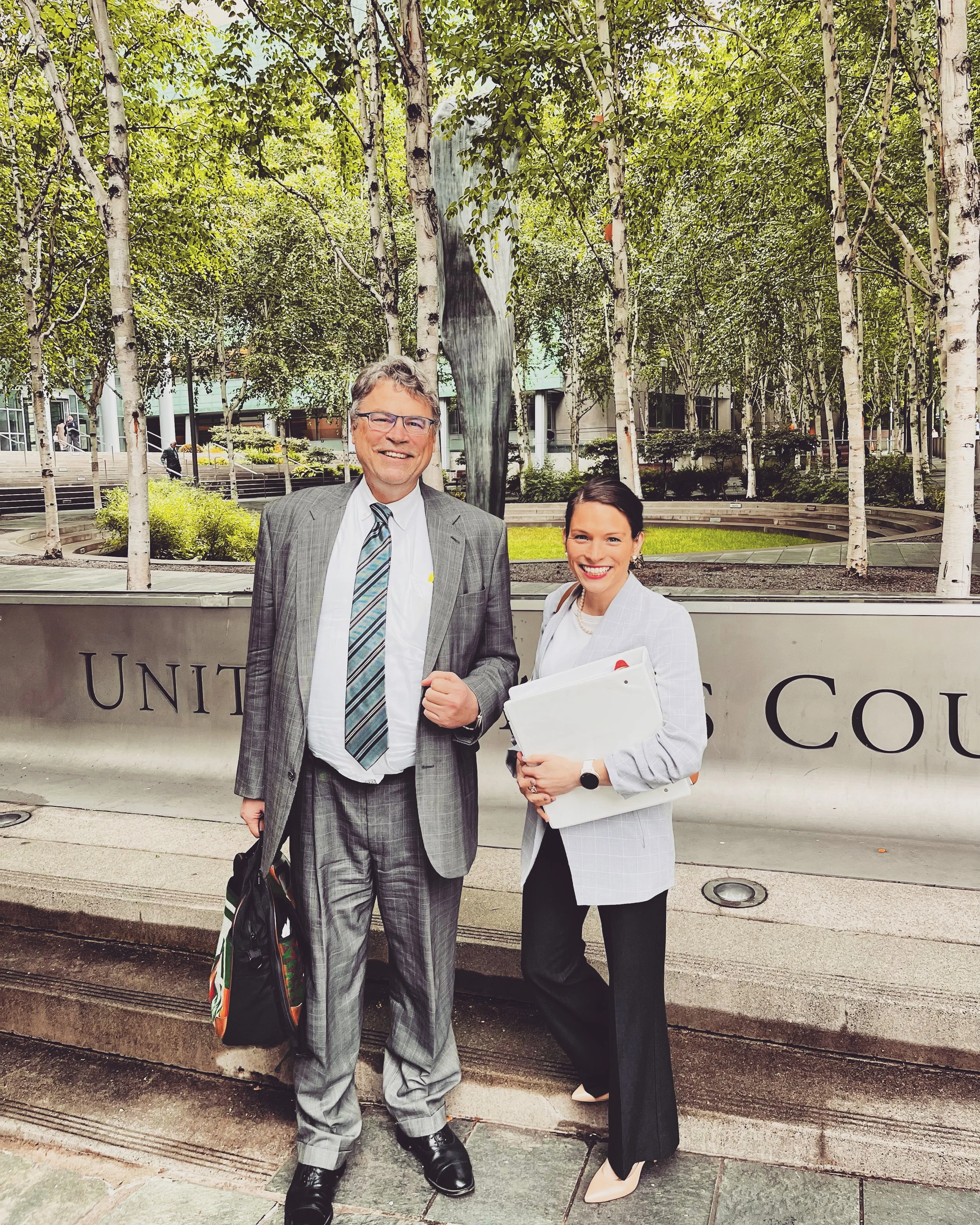 Two professionally dressed individuals, a man and a woman, standing outside in front of a concrete wall with trees and a sculpture in the background. The man is wearing a gray suit with a tie, and the woman is wearing a light gray blazer with black pants and holding papers.