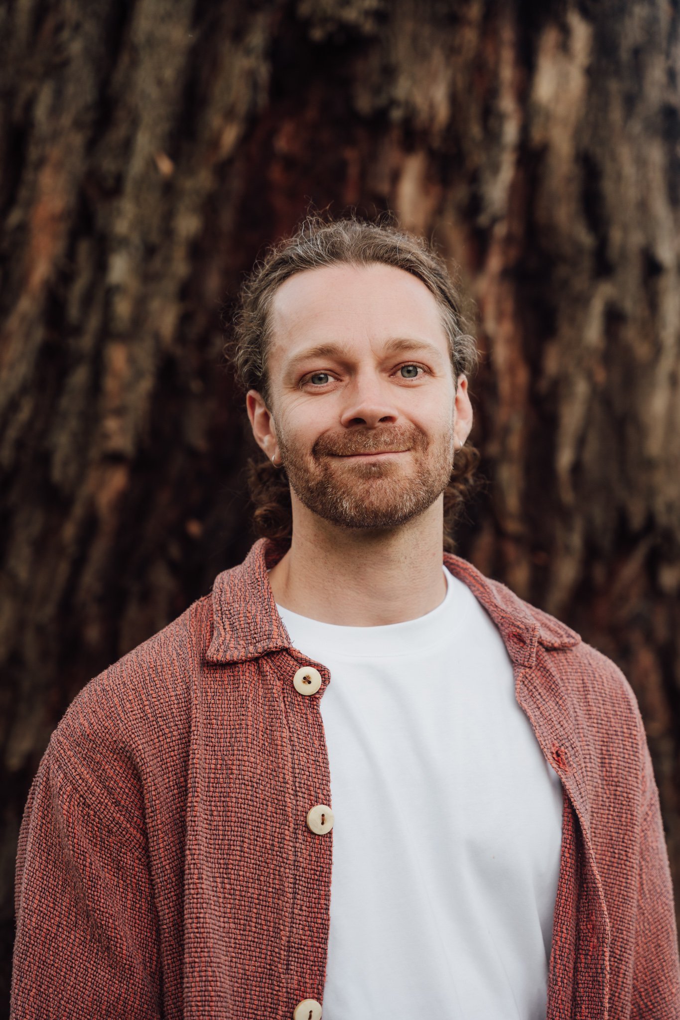 A man with light brown hair and a beard standing outdoors in front of a large tree.
