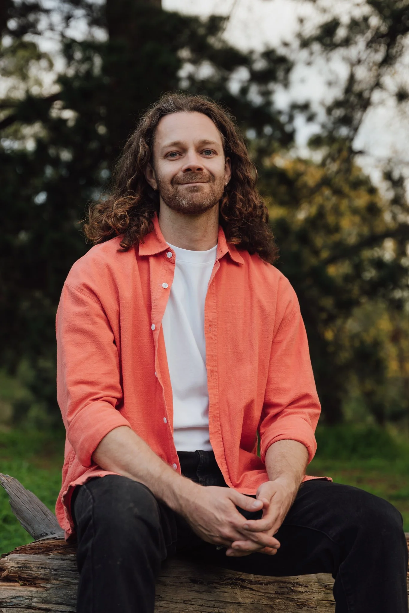 A man with long curly hair, a beard, and light skin sitting on a fallen log outdoors. He is wearing a coral button-up shirt over a white T-shirt and black pants, with a forest background of trees and foliage.