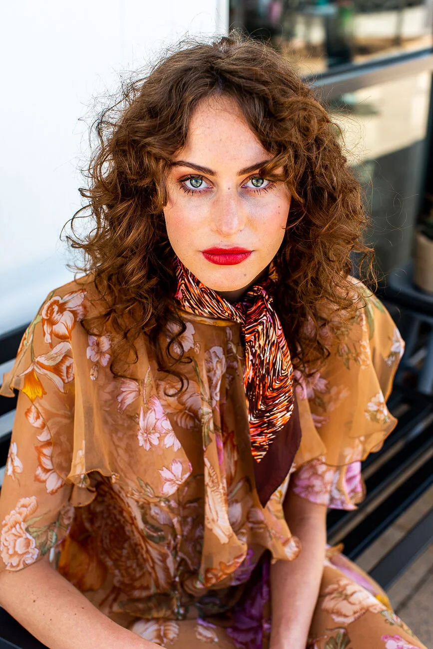 A young woman with curly brown hair and blue eyes, wearing a floral dress and a patterned scarf, seated outdoors with a blurred background.