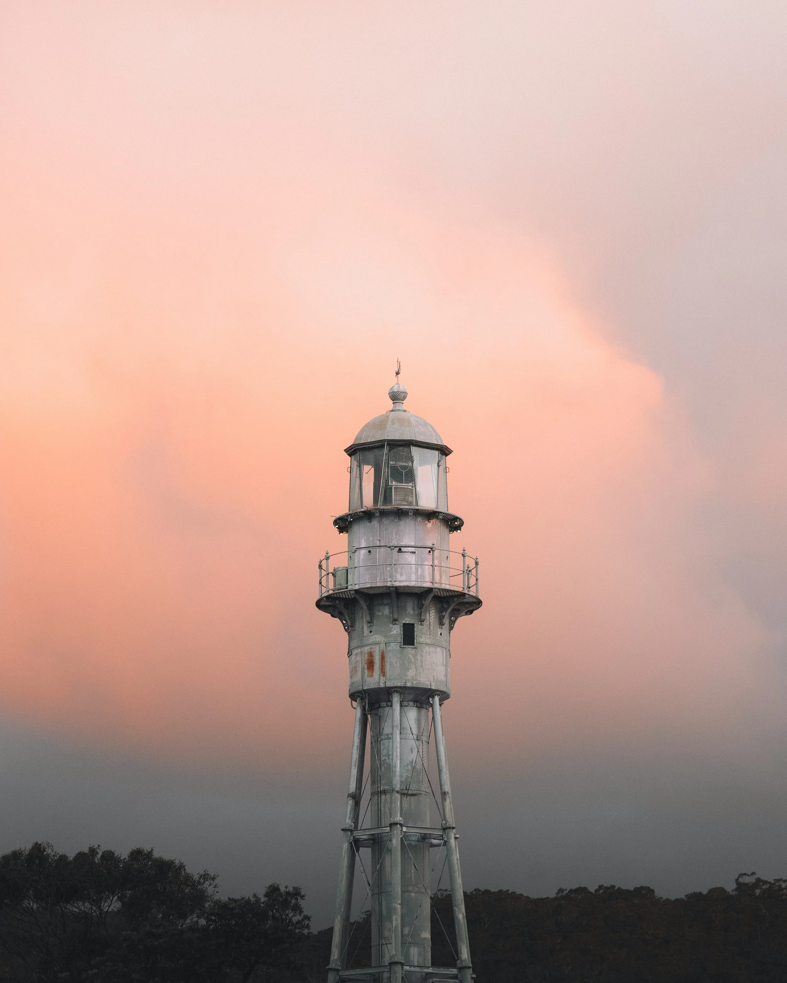 A lighthouse with a metal structure against a colorful sunset sky with pink, orange, and gray hues, surrounded by silhouette trees.