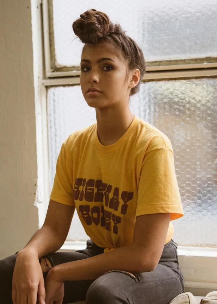 A young woman with brown hair styled in a bun, sitting by a textured glass window, wearing a yellow T-shirt with printed text and gray ripped jeans.