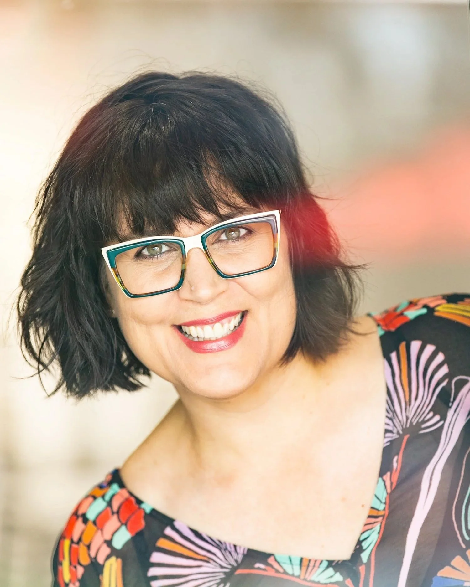 A smiling woman with short, wavy black hair wearing fashion glasses and a colorful patterned dress.
