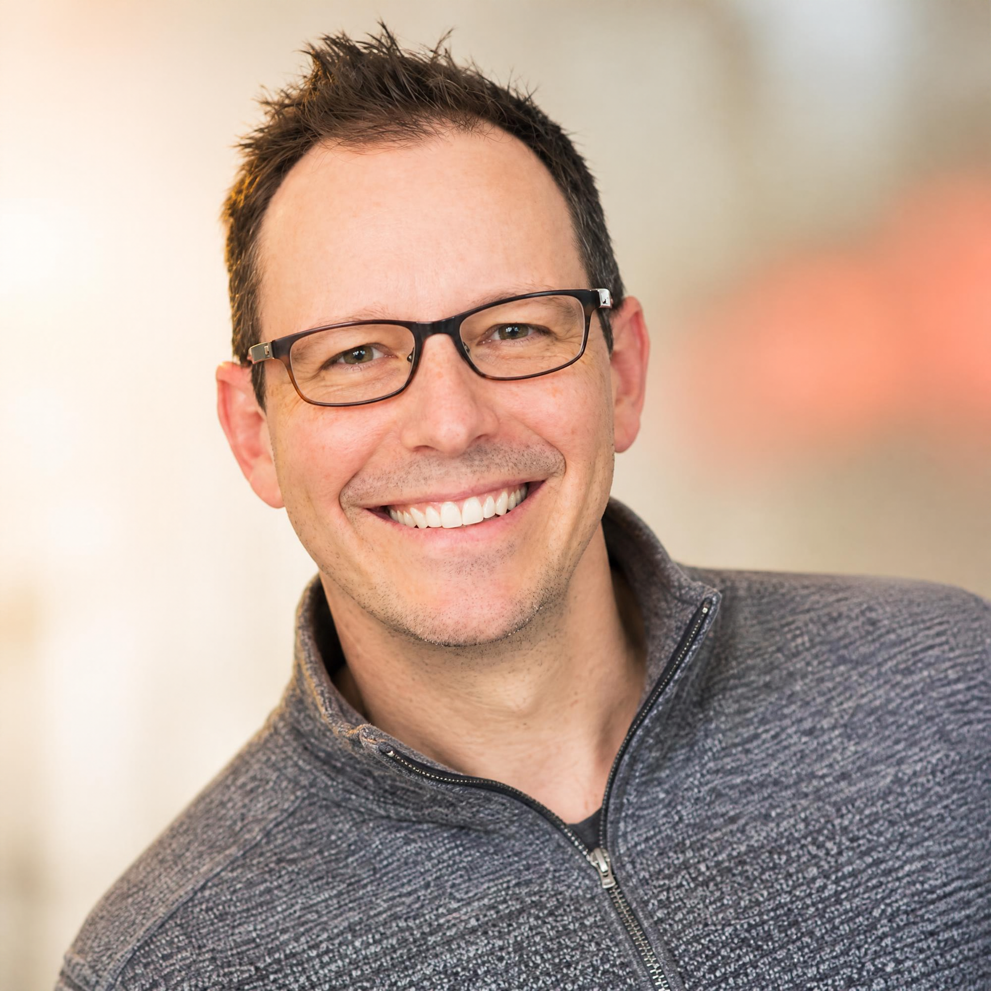 Partial view of a smiling man with glasses, short dark hair, wearing a gray zip-up sweater.