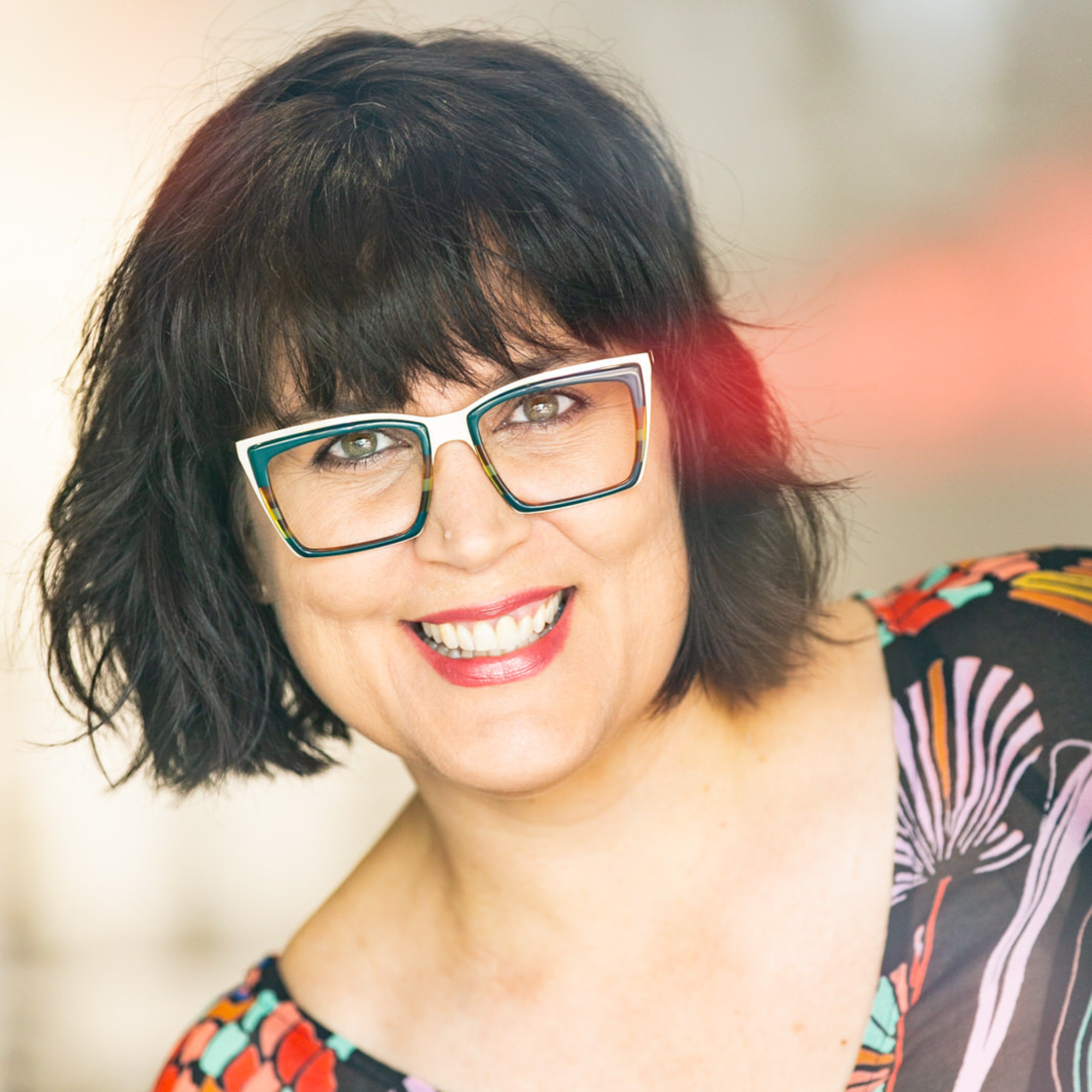 Close-up of a smiling woman with short black hair wearing colorful glasses and a multicolored striped shirt, against a plain background.