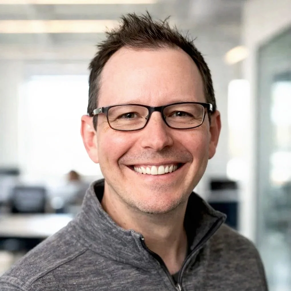 A smiling man wearing glasses and a gray zip-up jacket, in an office setting with blurred background.