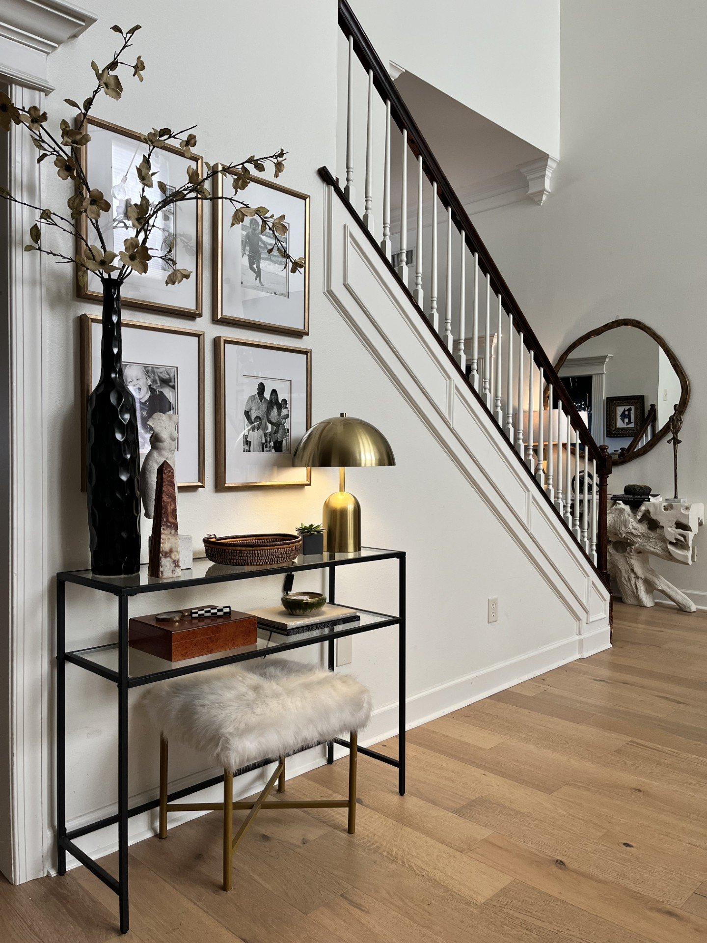 Interior of a modern home with a staircase, framed black and white photos on the wall, a black metal console table with decorative objects, a gold table lamp, a furry stool, and an arched wooden mirror.