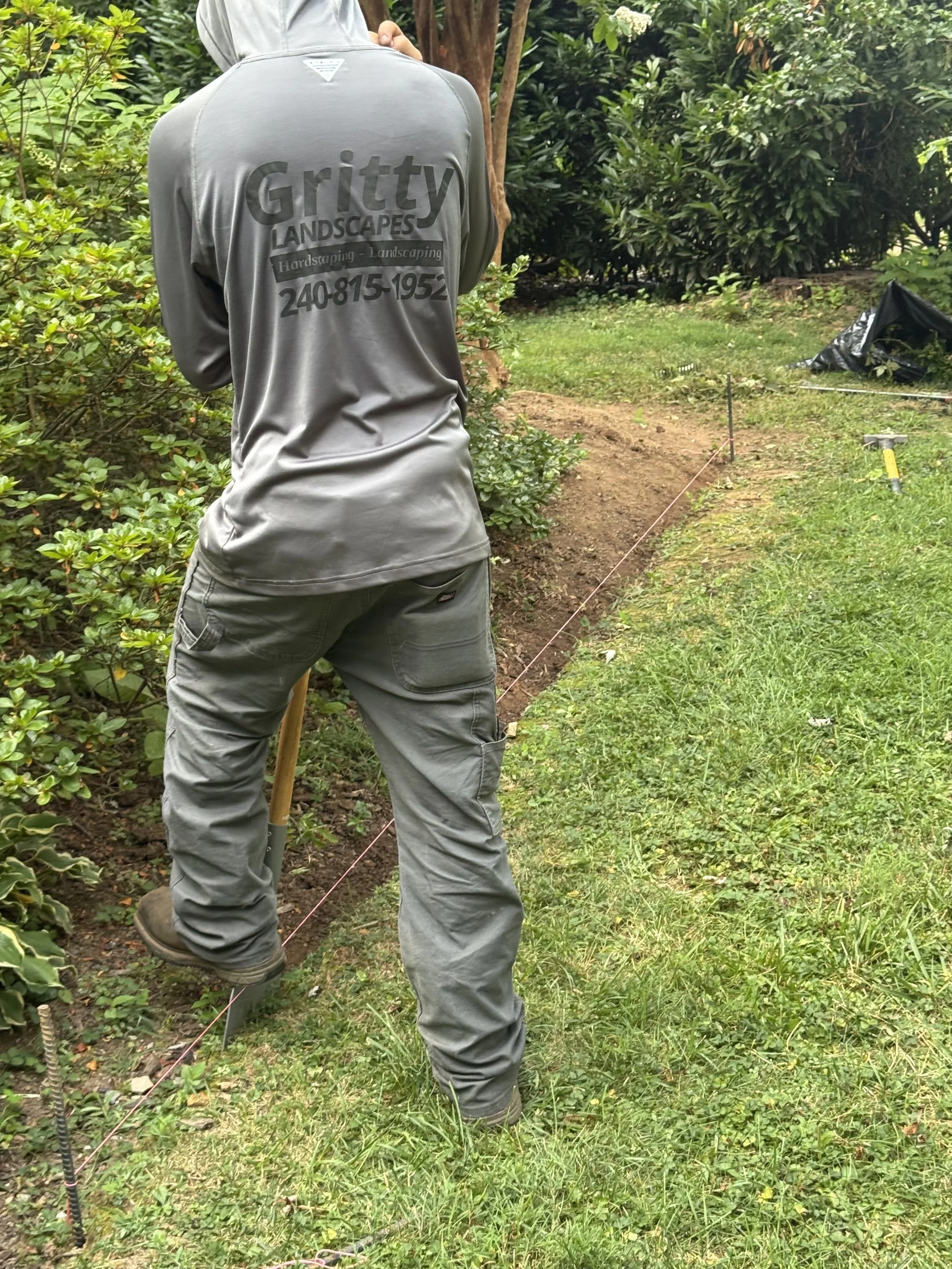 Person wearing a gray Gritty Landscapes jacket and cargo pants working outdoors, with surveying equipment and a marked boundary line on the ground, surrounded by green bushes and trees.
