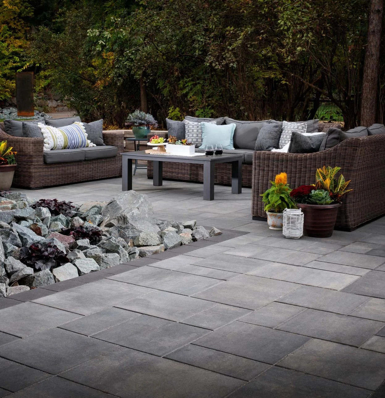 Outdoor patio with gray stone flooring, wicker sofas with cushions, potted plants, and a black coffee table with drinks and a fruit tray, surrounded by green trees.