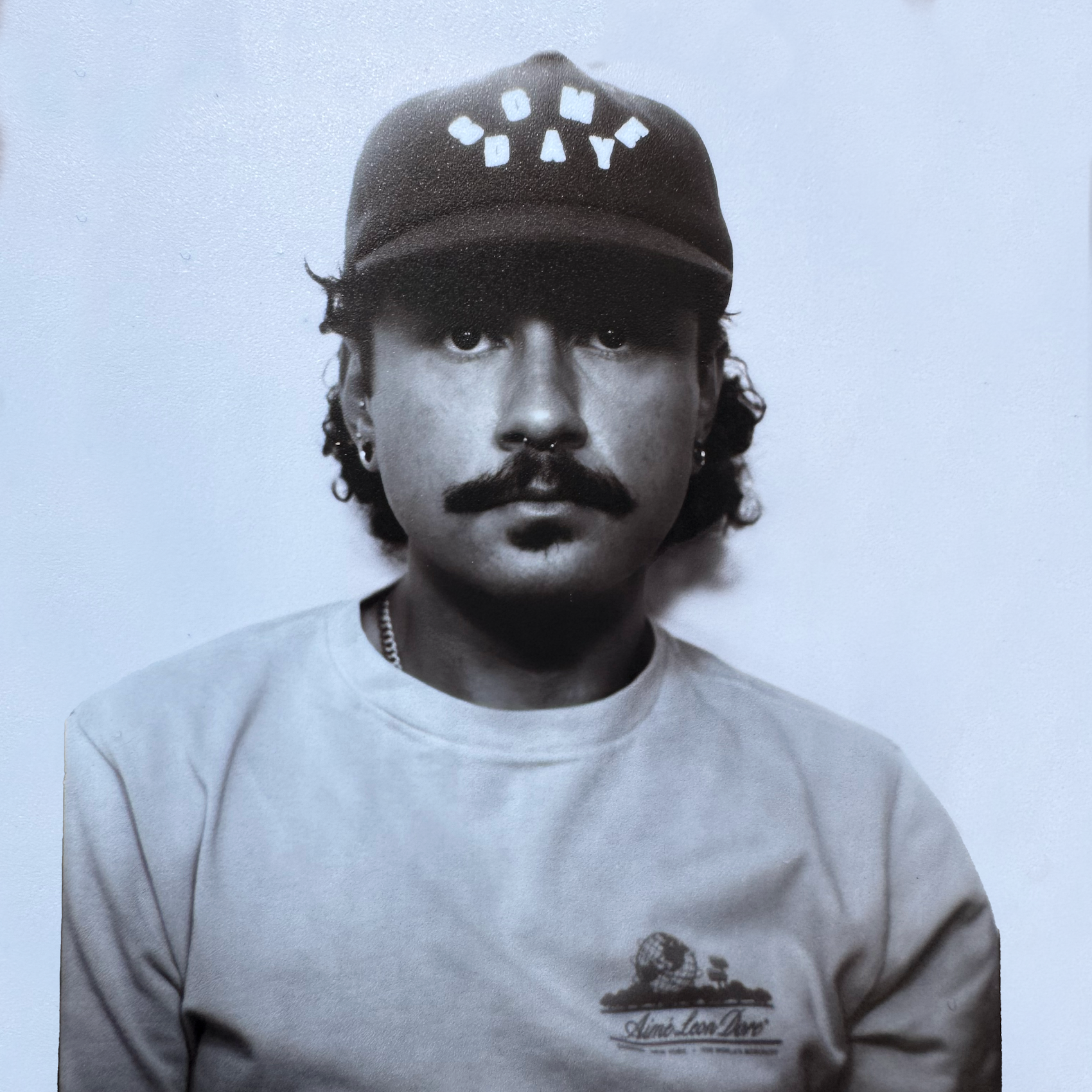 Black and white portrait of Phillip Antonio Lemos with a moustache and curly hair wearing a baseball cap with the word "DAY" on it, a nose ring, earrings, a chain necklace, and a T-shirt with a graphic design and text.