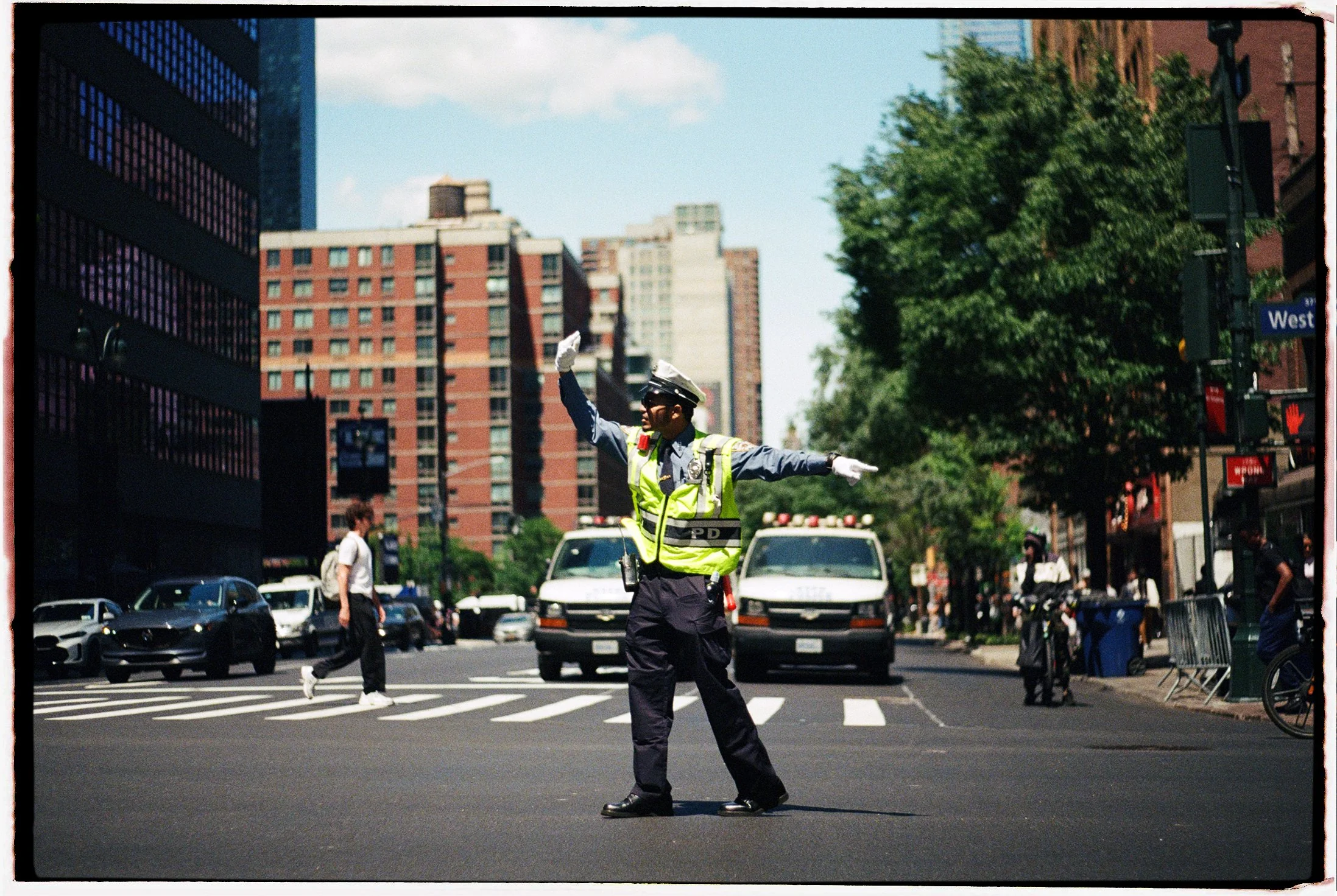 A police officer standing in the middle of a New York city street, directing traffic with arms outstretched. The scene includes tall buildings, pedestrians, and cars.