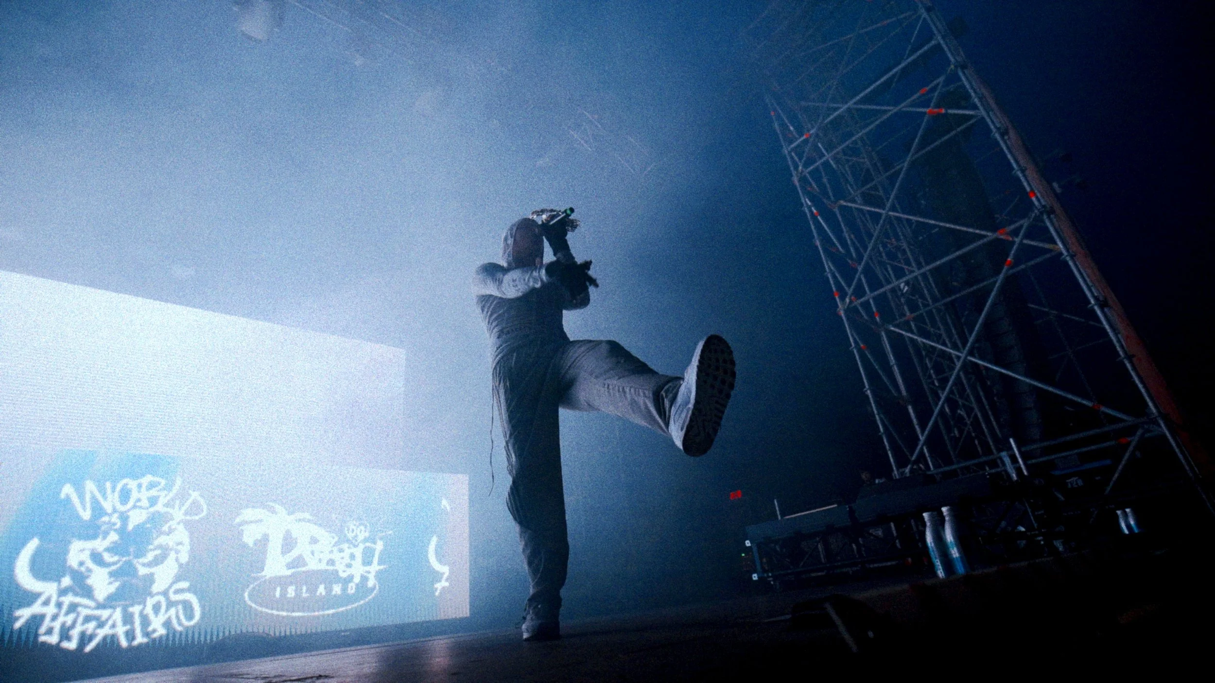 Young Lean on stage at Laneway Sydney 2025 with one leg raised, wearing gray pants and a black jacket, holding a microphone, near a metal scaffold structure, with a large screen or projection in the background displaying graffiti-style text.
