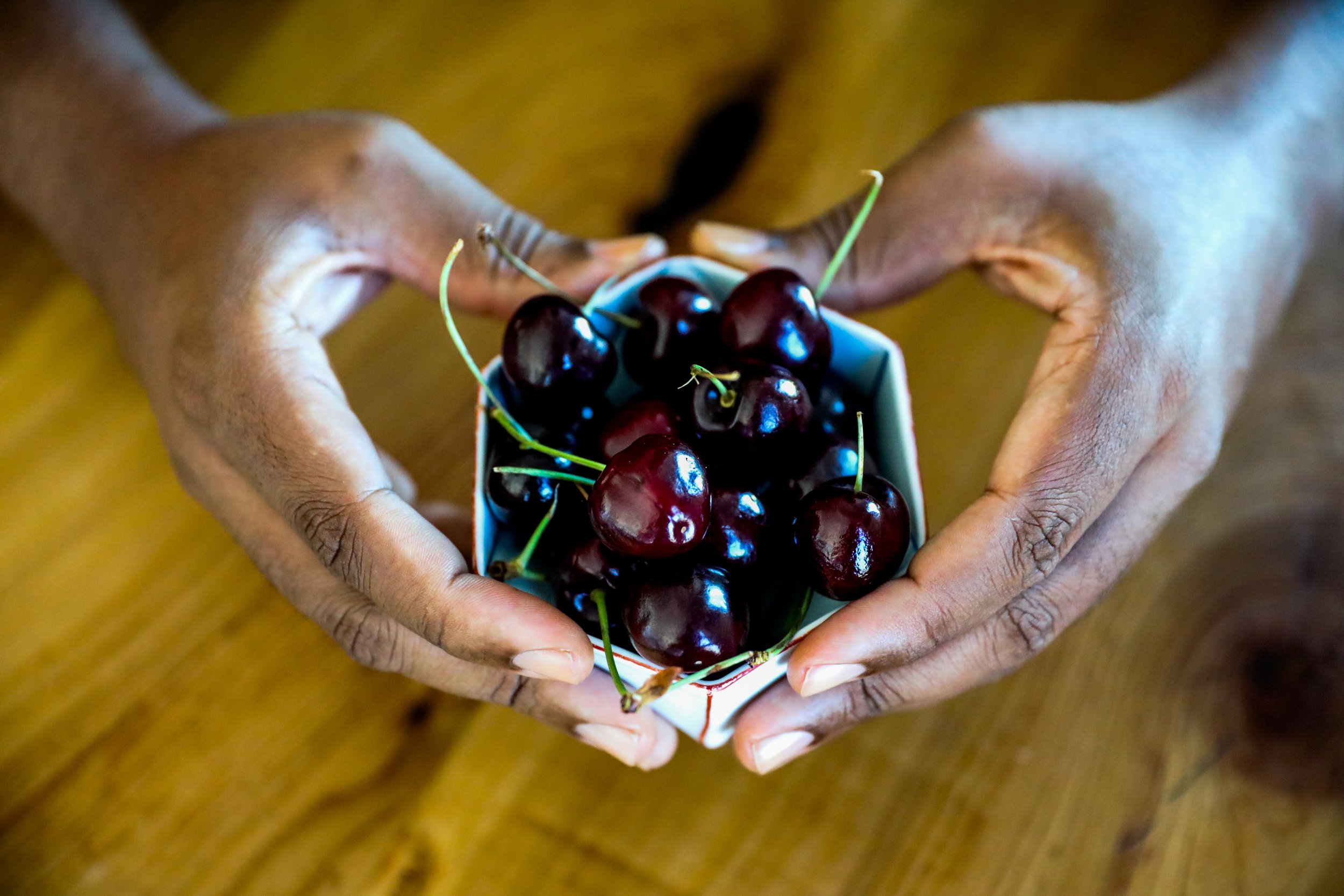 Person with a richly hued, medium dark skin tone and clean, clear nailbeds, whose hands form the shape of a heart, holding a small clay origami shaped cup filled with dark red cherries over a wooden surface. 