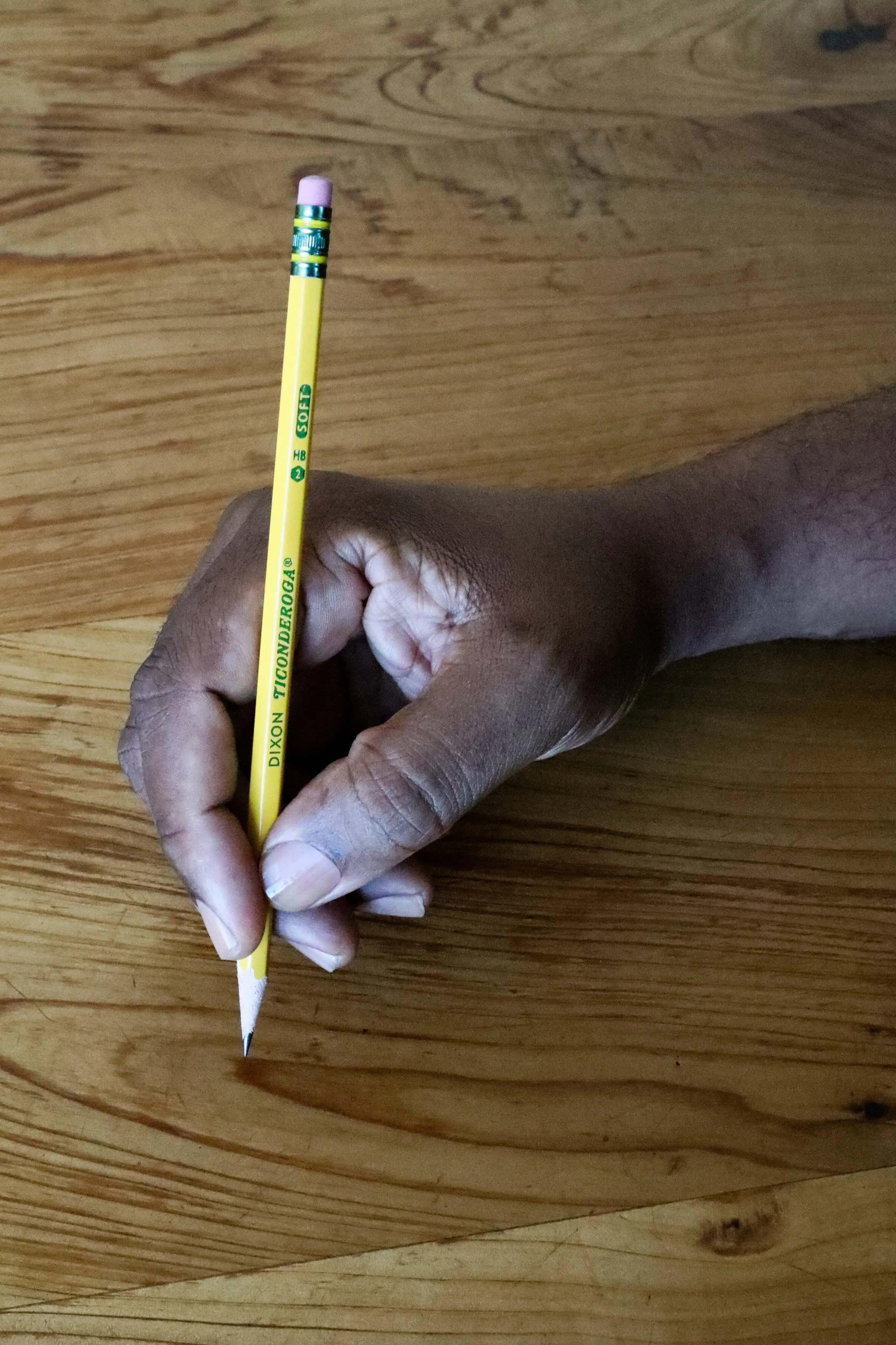 A richly hued, medium dark skinned hand with clean, clear nailbeds holding a yellow Dixon Ticonderoga No. 2 soft graphite pencil with pink eraser positioned over a wooden surface.