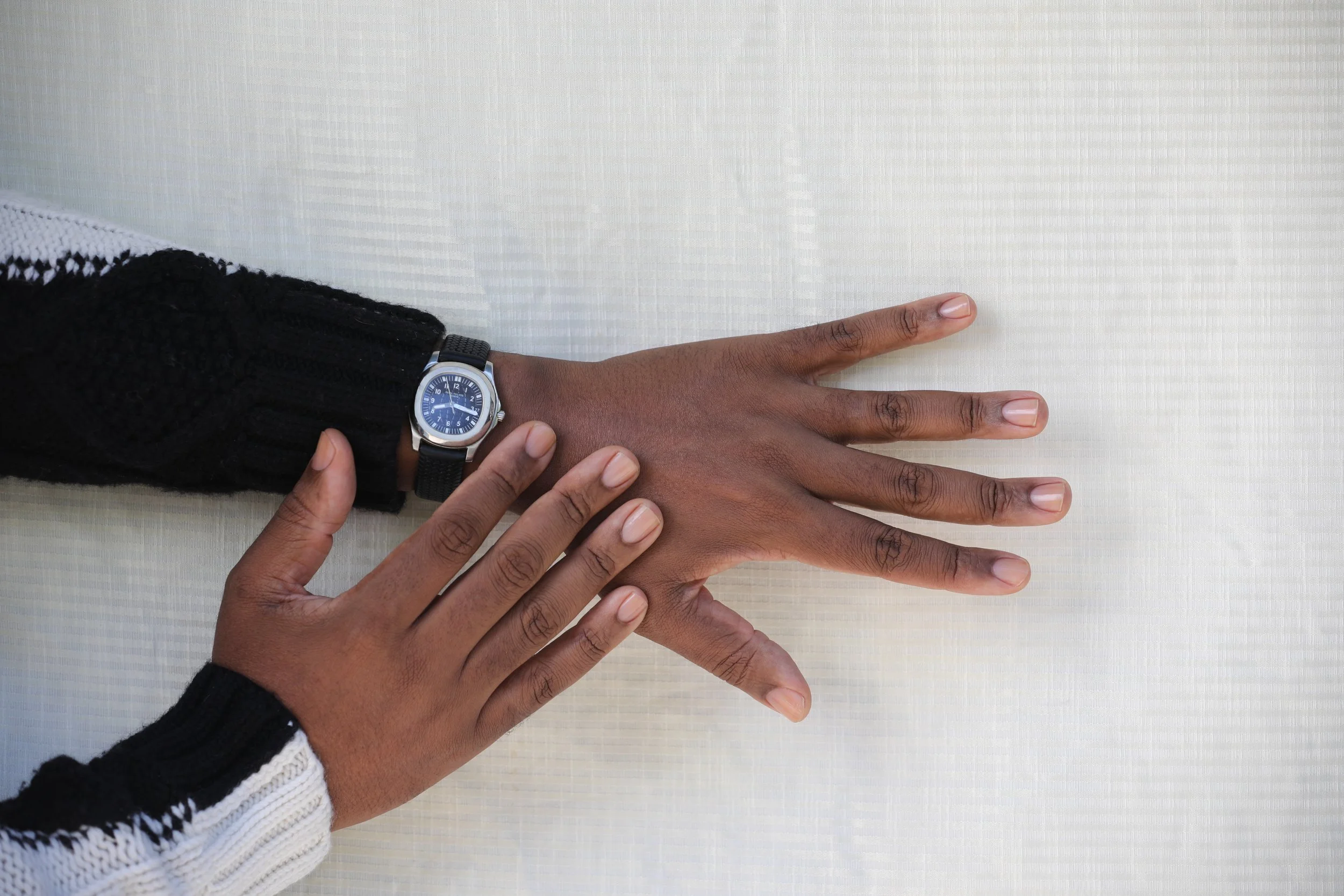 Person with richly hued, medium dark skin tone and clean, clear nailbeds wearing a black and silver leather wristwatch and black and white wool sweater, holding out their left and right hands against a white surface.
