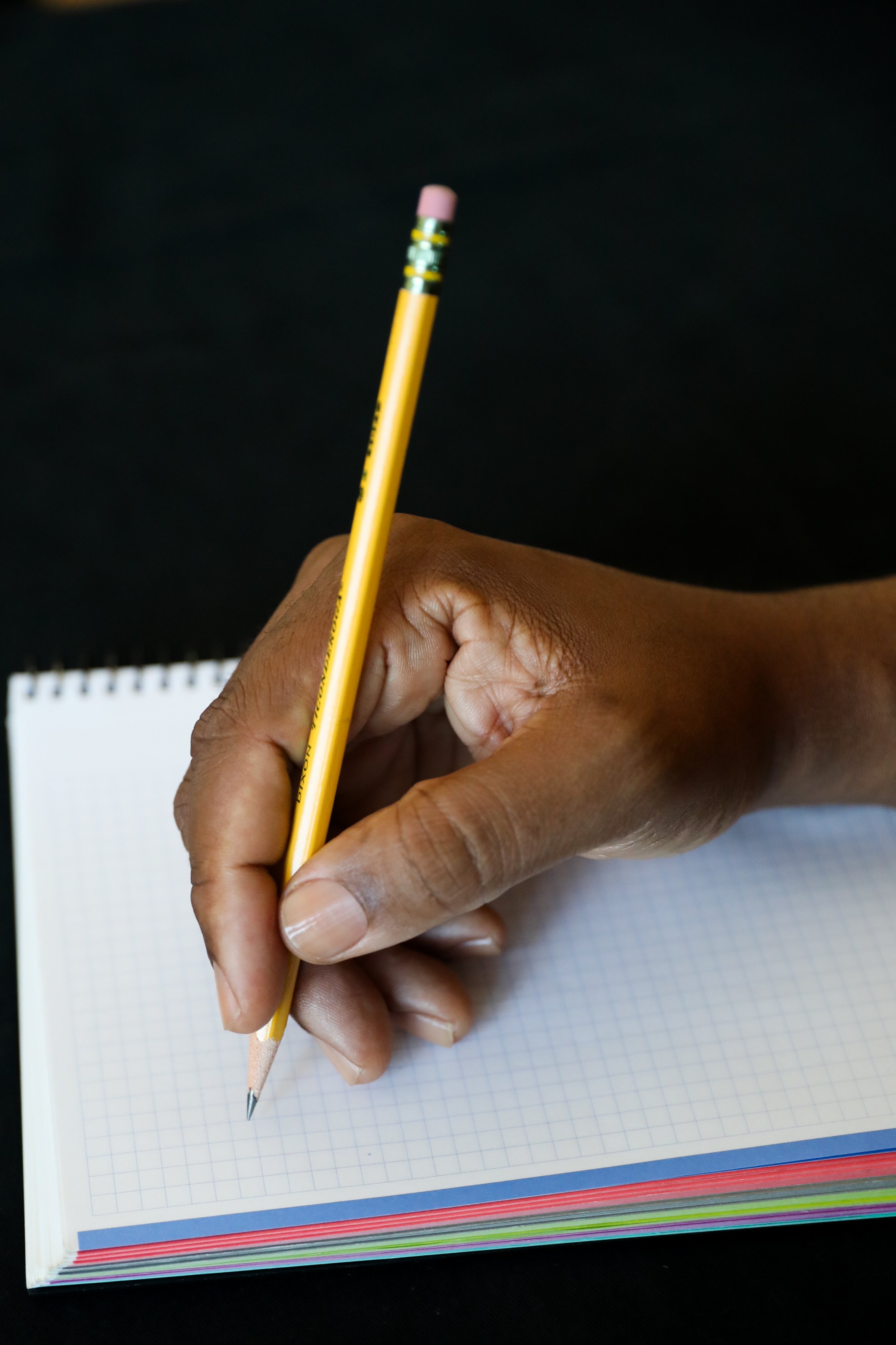 A richly hued,  medium dark skin tone right hand with clean and clear nailbeds lholding a sharpened yellow pencil with a pink eraser, poised above an open graph paper notebook with multicolored pages underneath, on a black background.