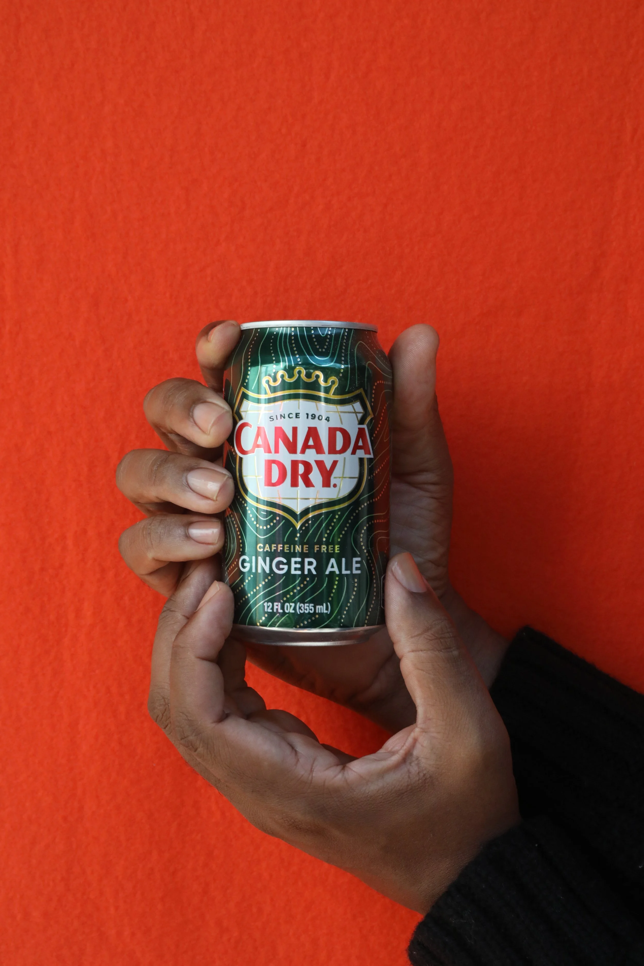 Two richly hued, medium dark skin tone hands with clean, clear nailbeds holding a can of Canada Dry Ginger Ale against a bright orange background.