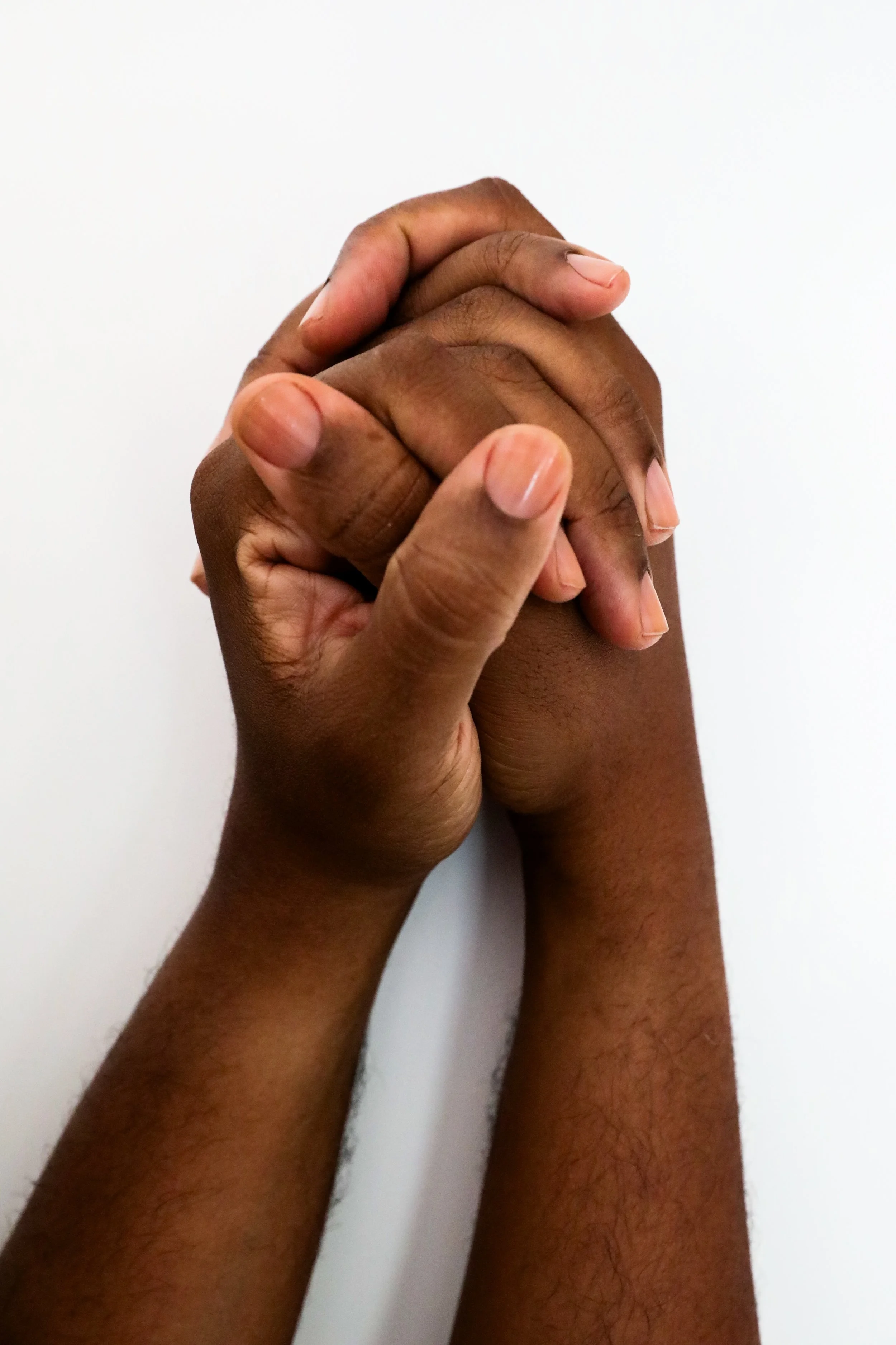 A close-up of two richly hued, medium dark hands with clean, clear nailbeds clasped together against a plain white background.