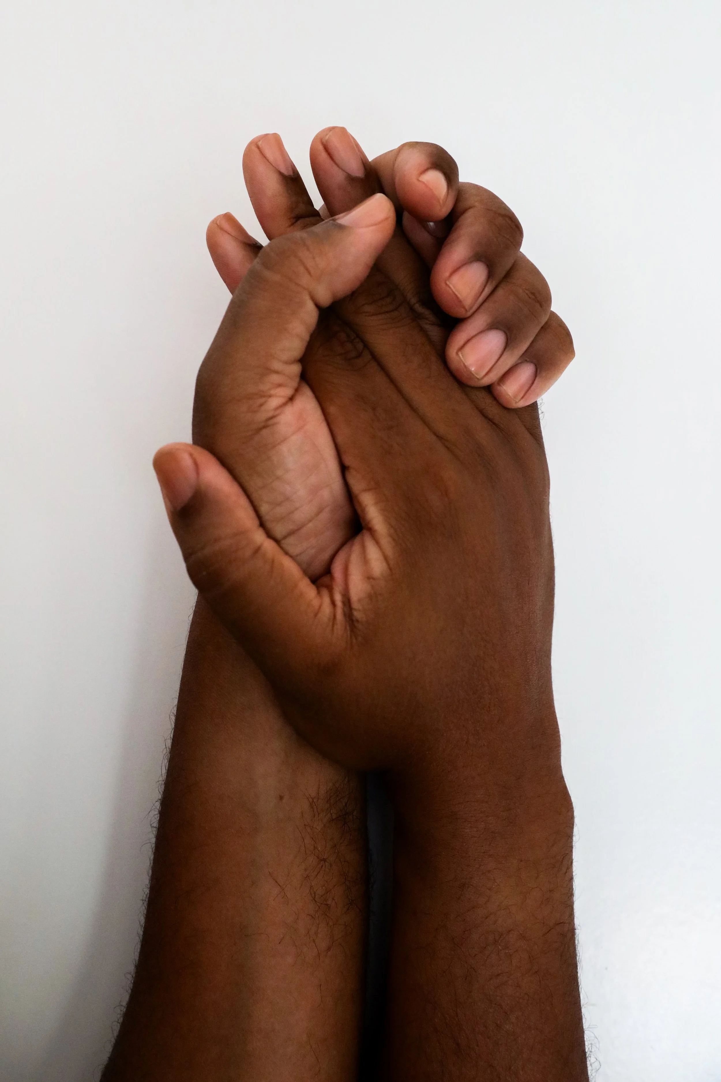 Two richly hued, medium dark hands with clean, clear nailbeds holding each other against a white background.