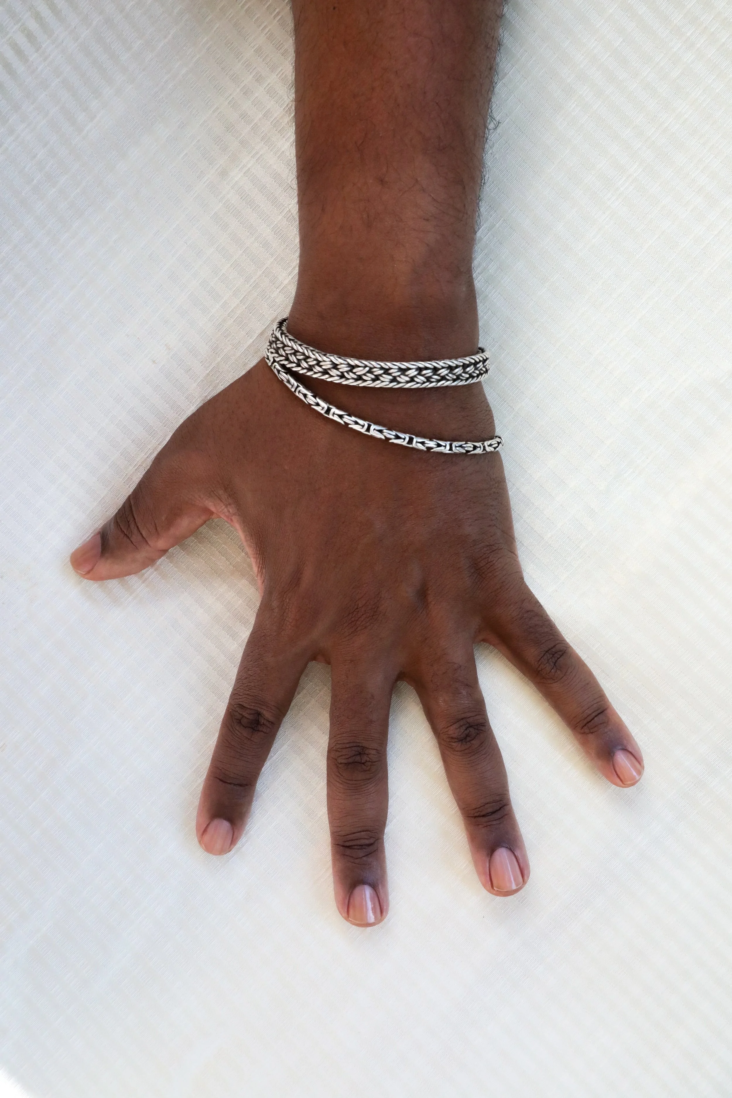 A richly hued, medium dark skin tone with clean, clear nailbeds left hand wearing two silver bracelets resting on a white textured surface.
