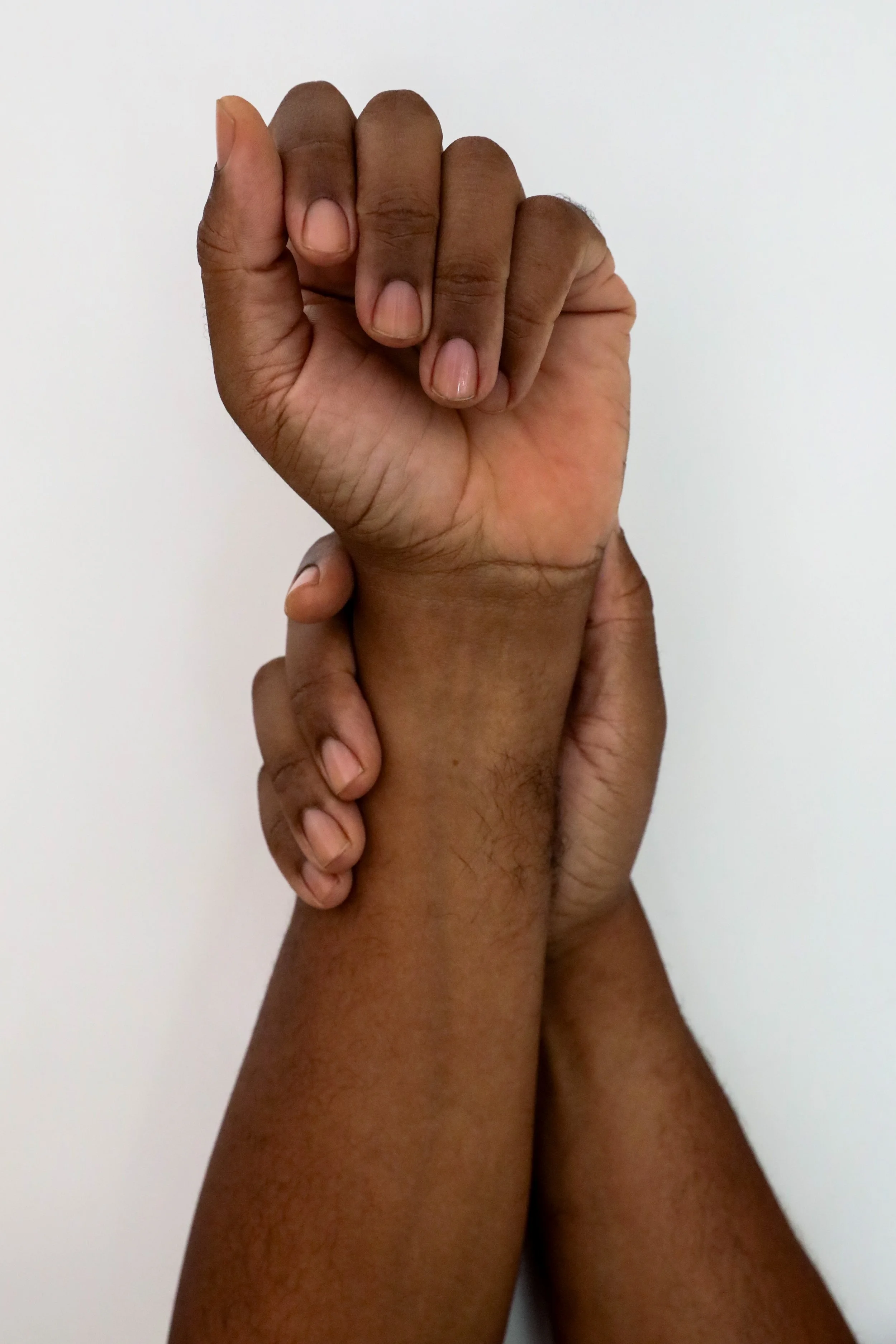 Two richly hued, medium dark hands with clean, clear nailbeds, one grasping the other, against a plain light background.