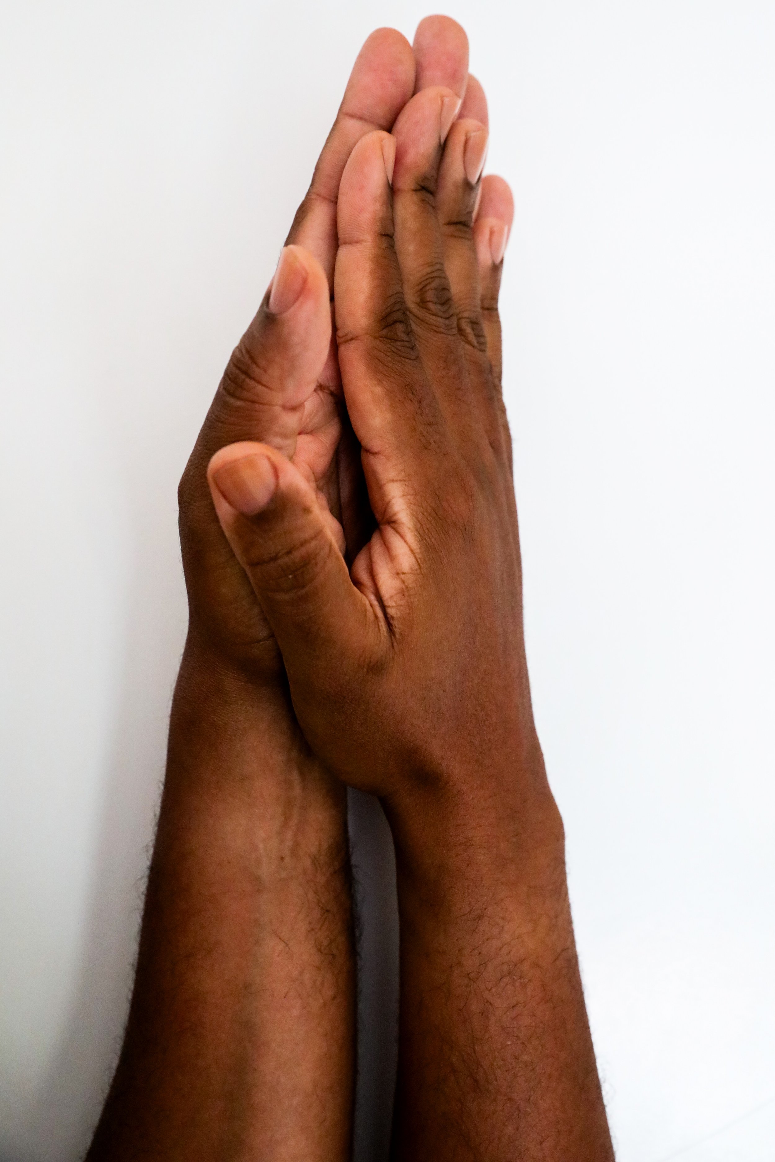 Two hands with medium dark, rich skin tone and clean, clear nailbeds artistically pressed together against a plain white background.
